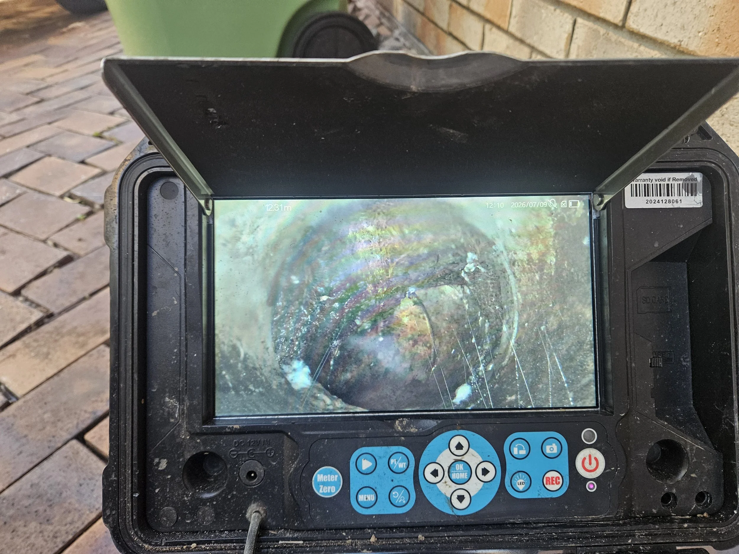 A handheld electronic device with a cracked and smudged screen, showing a rainbow oil film or bubble on water surface, placed on a table outdoors near a brick wall and a green trash bin.