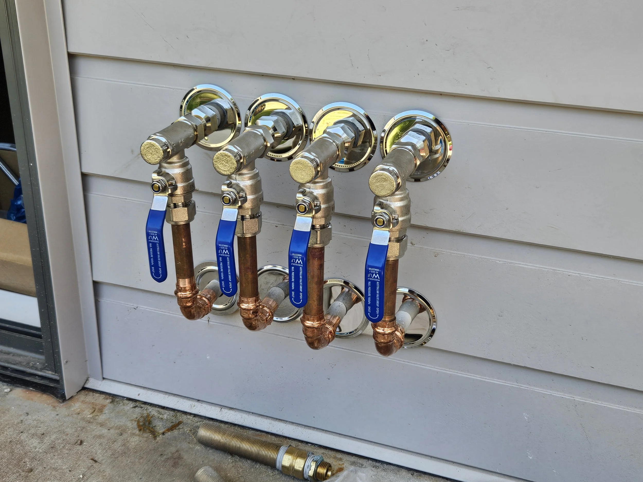 A plumbing setup with four copper pipes connected to a white wall, each pipe with a blue shut-off valve and a chrome connection, situated outdoors on a concrete surface.