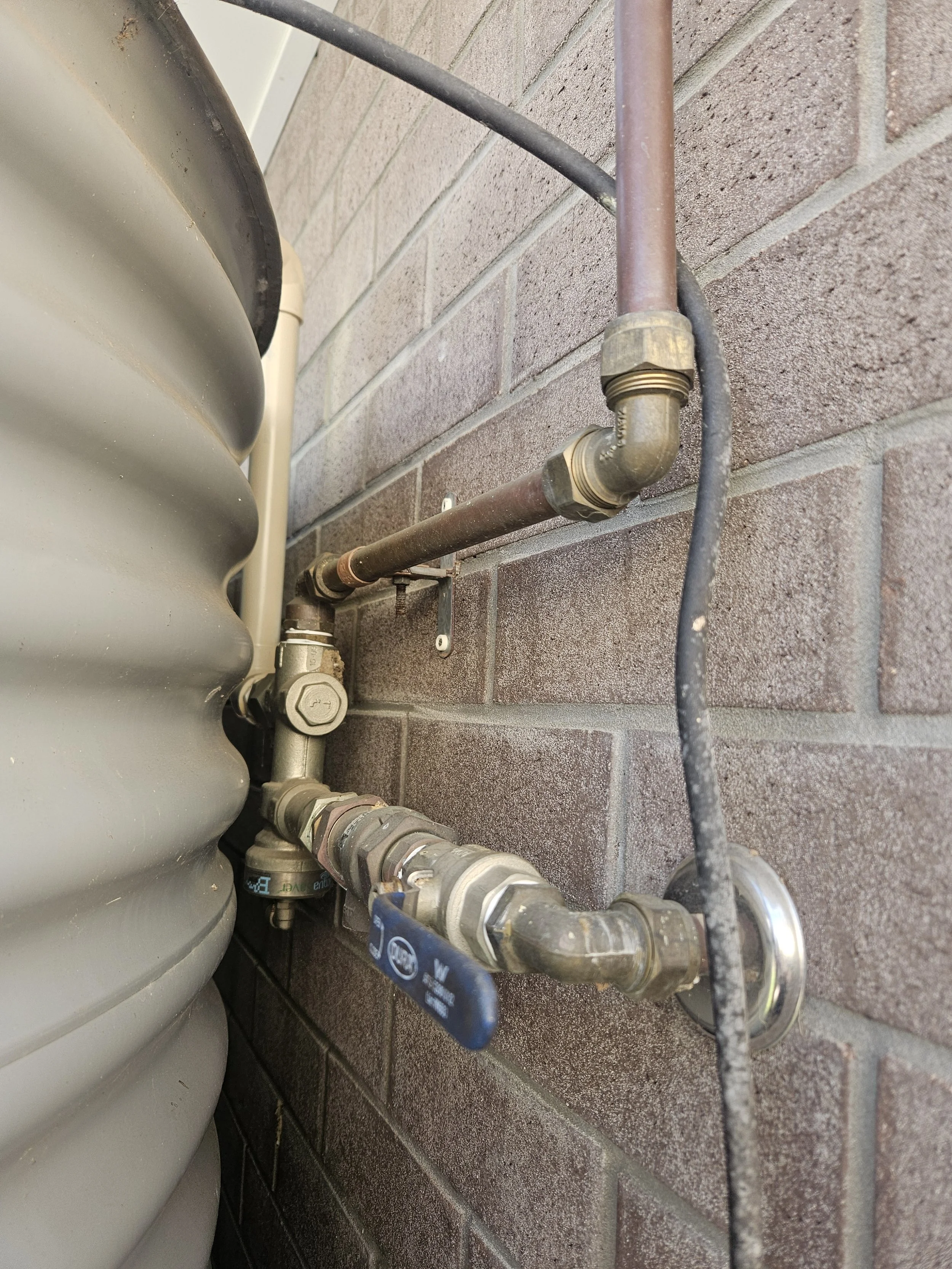 Close-up of plumbing pipes and fittings on a brick wall, with a section of an HVAC unit on the left.