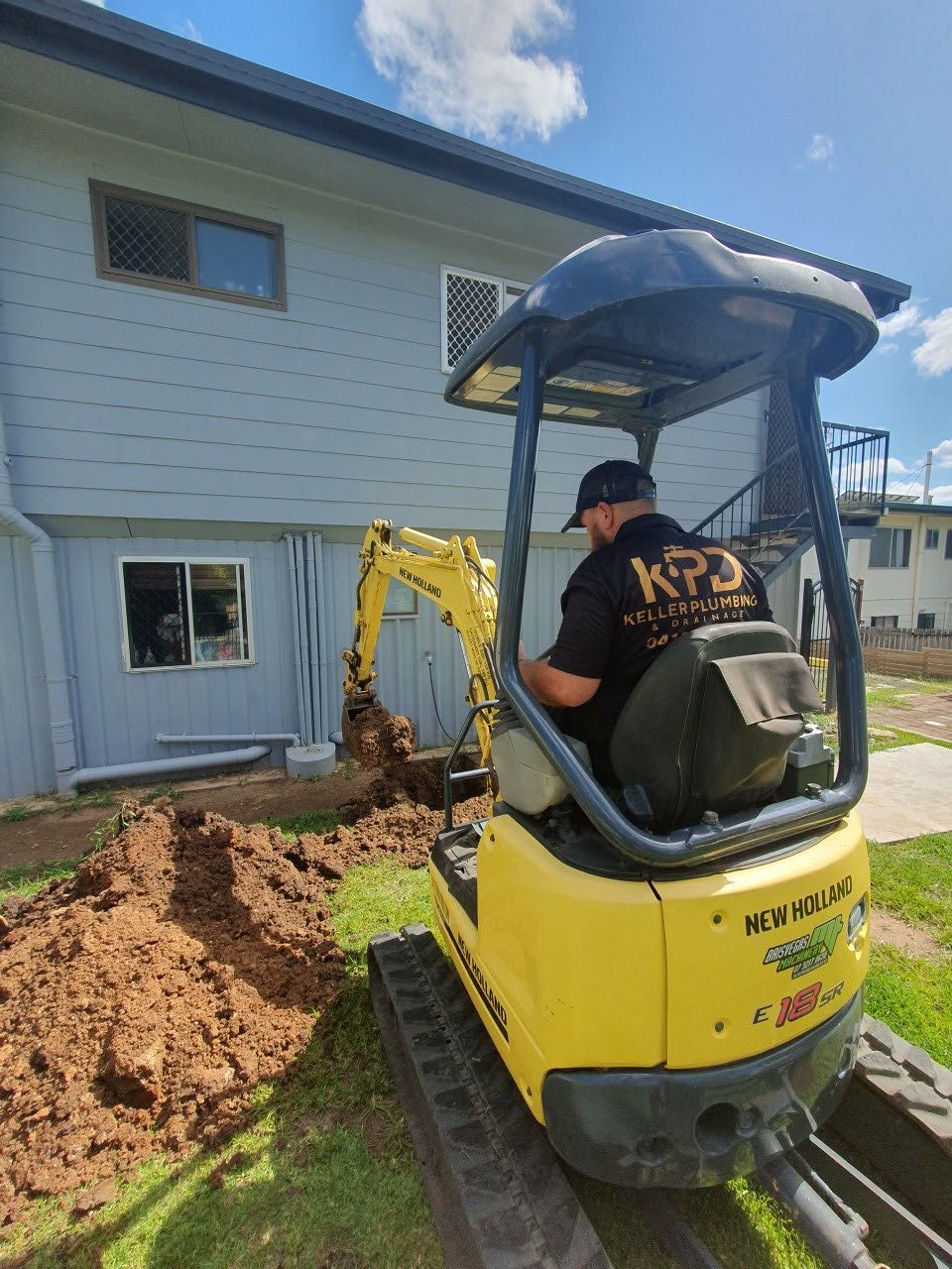 A worker operating a small yellow New Holland excavator digging a trench near the foundation of a house with light blue siding and white window frames, under a partly cloudy sky.