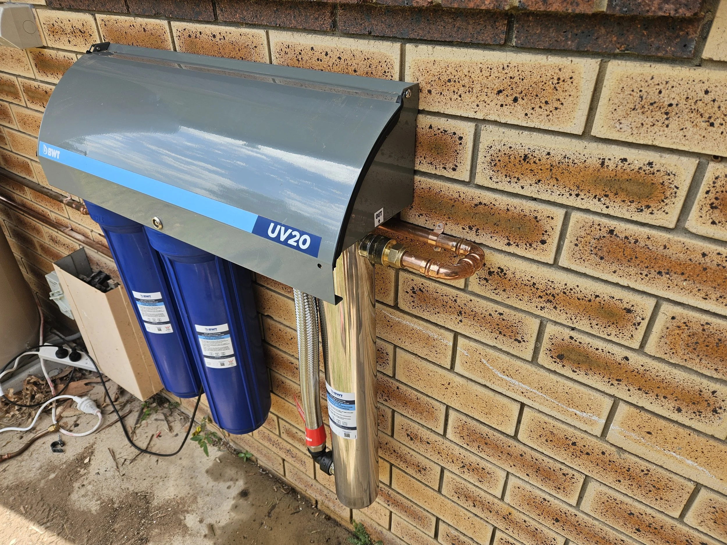 A water filtration system mounted on a brick wall outdoors, consisting of a blue water filter with a grey cover labeled "UV20" and pipes connected to it, with some electrical cords and ground nearby.