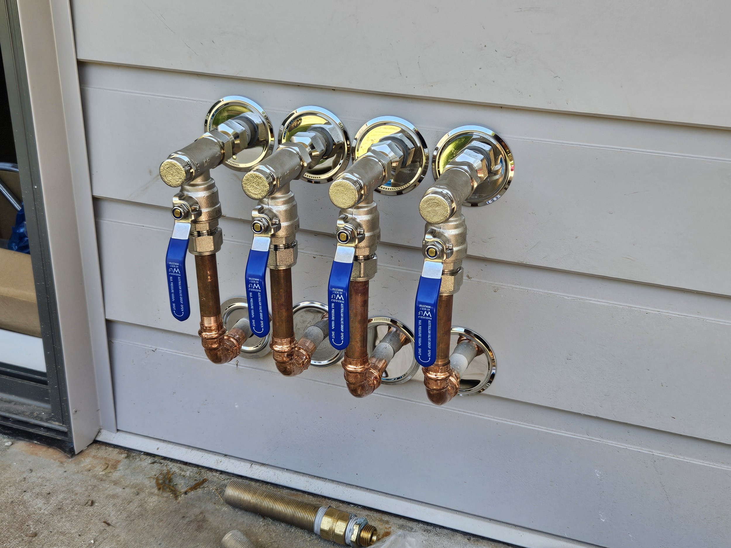 Four copper water supply pipes with shut-off valves and blue handles are installed on an exterior wall, with some flexible tubing and fittings on the ground nearby.