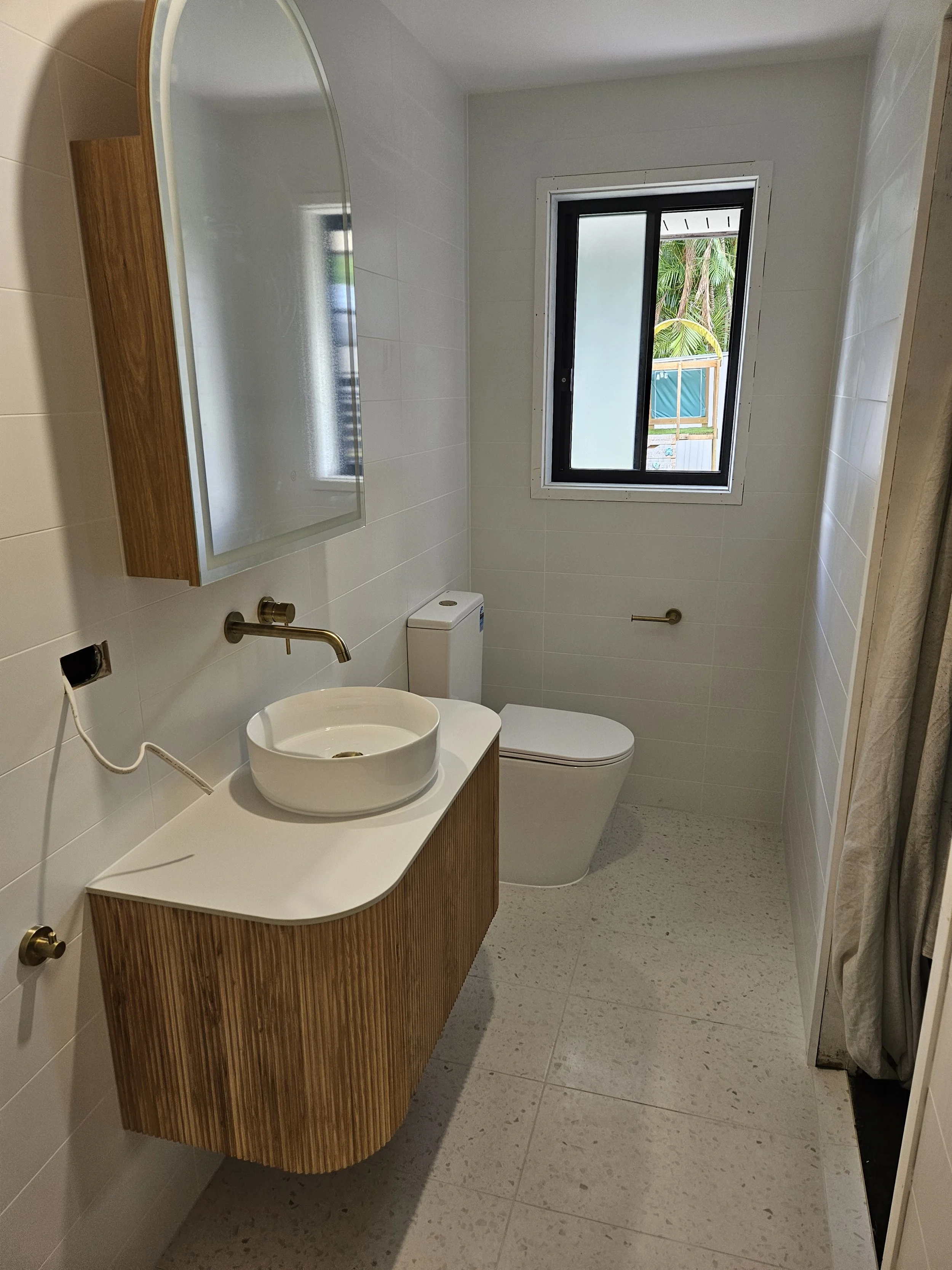 A modern bathroom with beige tiled floor and walls, featuring a wooden vanity with a vessel sink, a mirror above, a toilet, a nearby window showing outdoor greenery, and a shower area with a curtain.