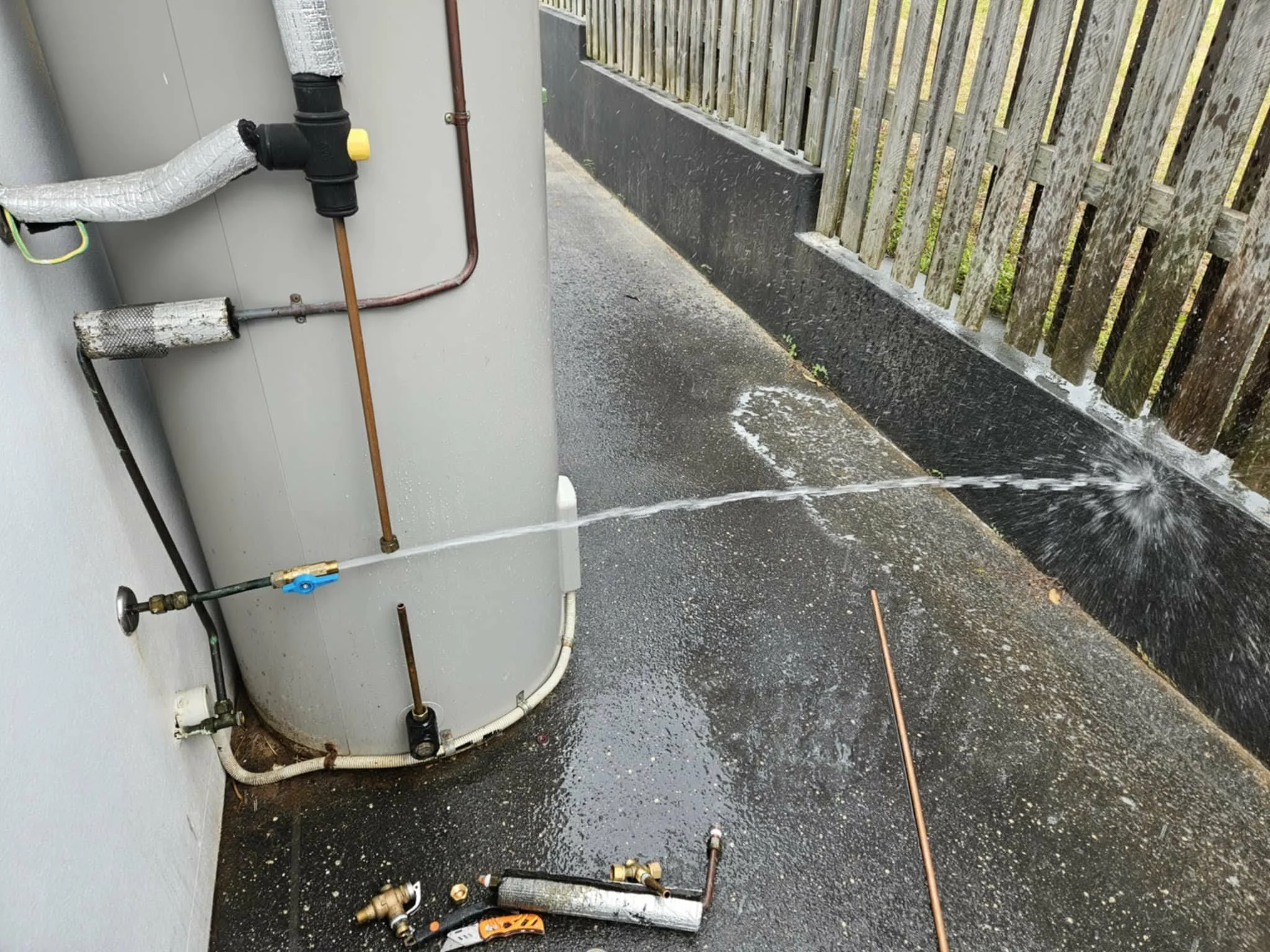 An outdoor water heater with pipes, valves, and insulation. Water is spraying on the black asphalt ground next to a wooden fence.