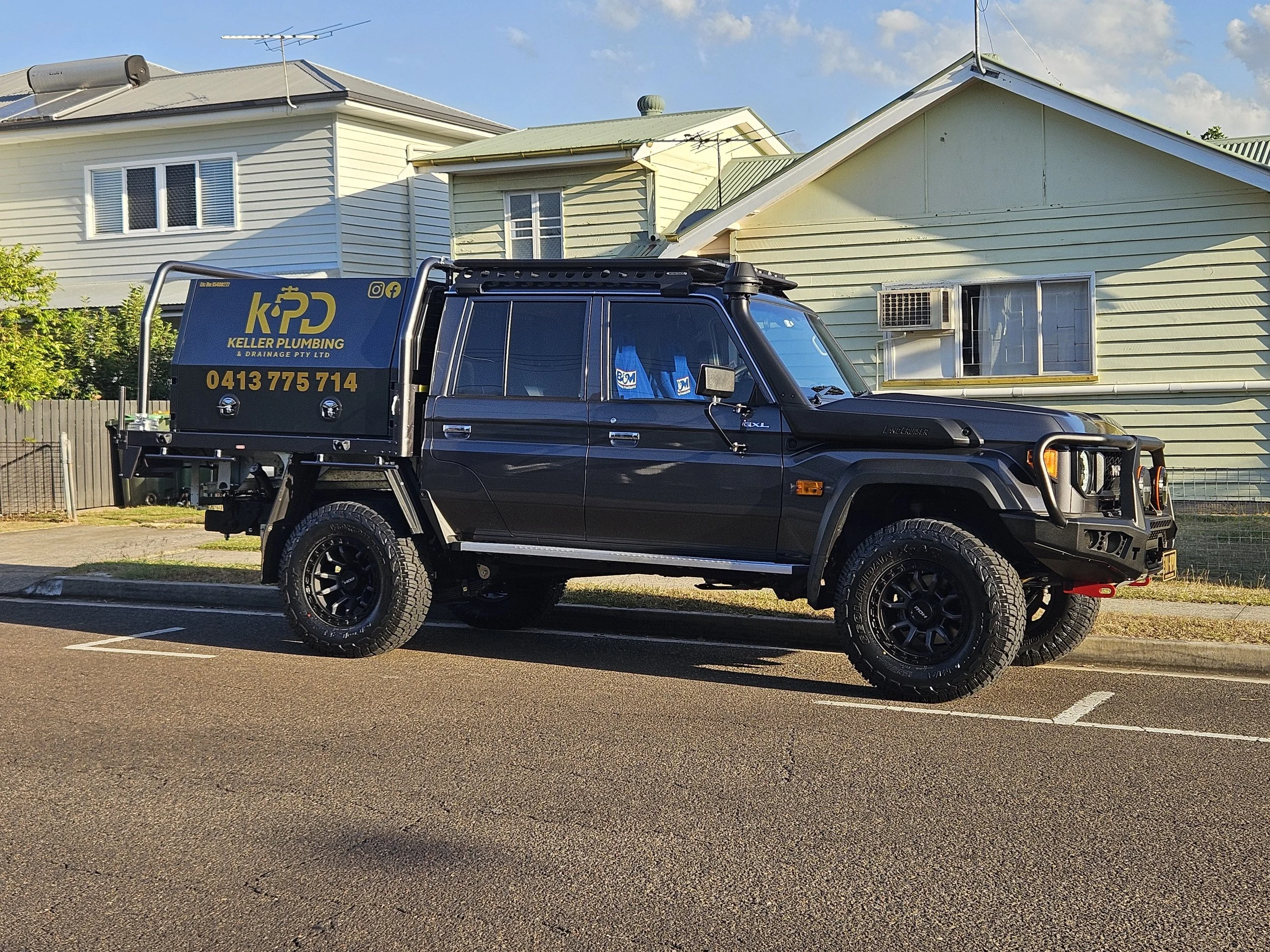 A black pickup truck parked in a parking lot with houses in the background. The truck has company branding for Keller Plumbing and Drainage, including a phone number and social media icons.