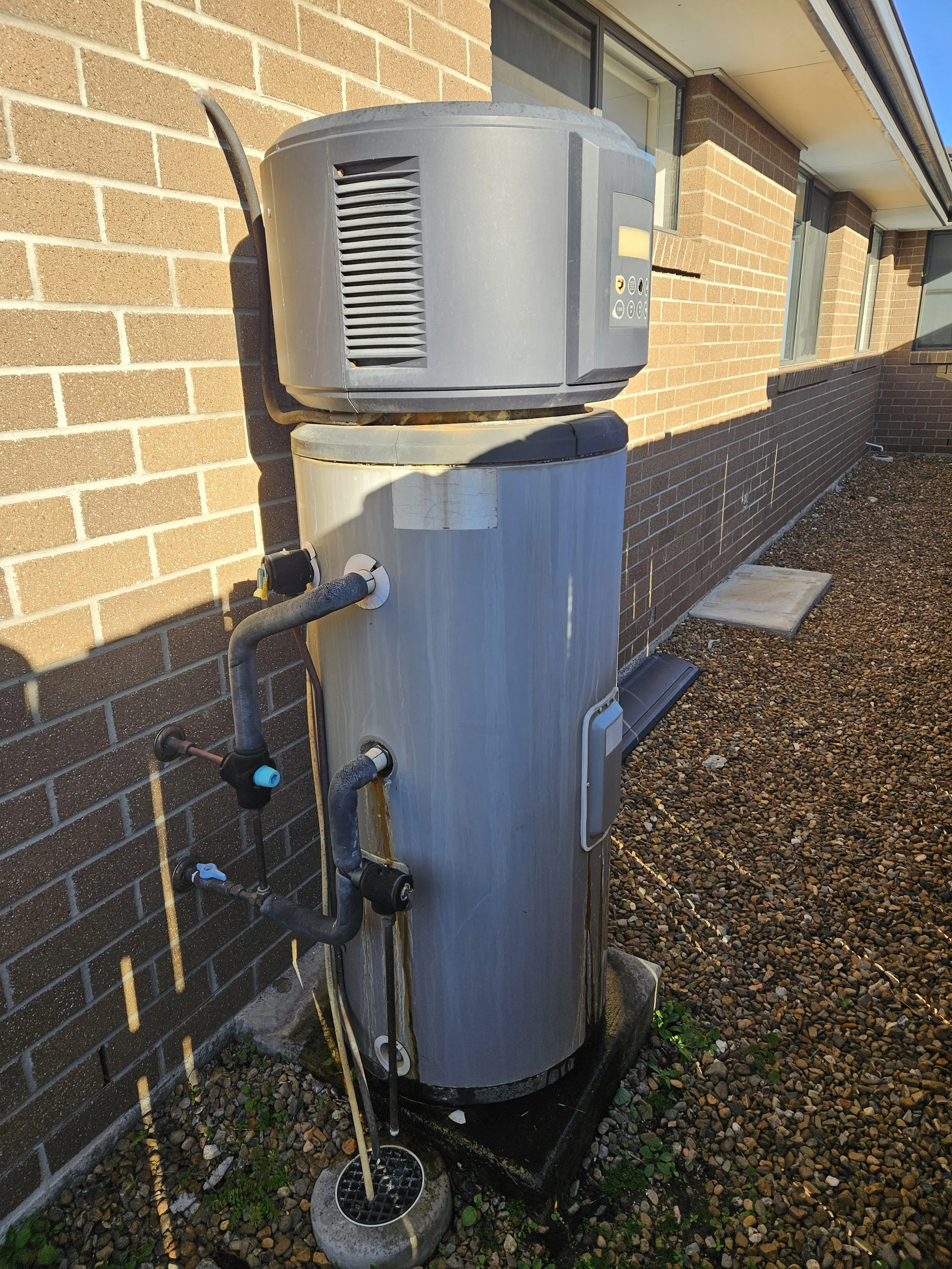 The image shows an outdoor air conditioning unit installed on a brick wall. The unit is mounted on a concrete pad and connected to pipes and electrical conduits. There is a drain pan at the bottom with some water and a garden trowel placed in it. The background features a gravel ground and a brick building with windows.