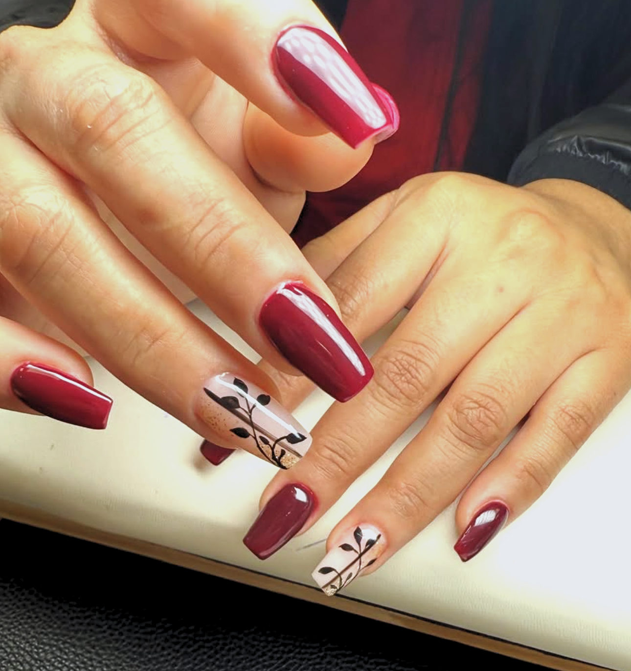 Close-up of hands with manicured nails painted in dark red, featuring black leaf designs on some nails.