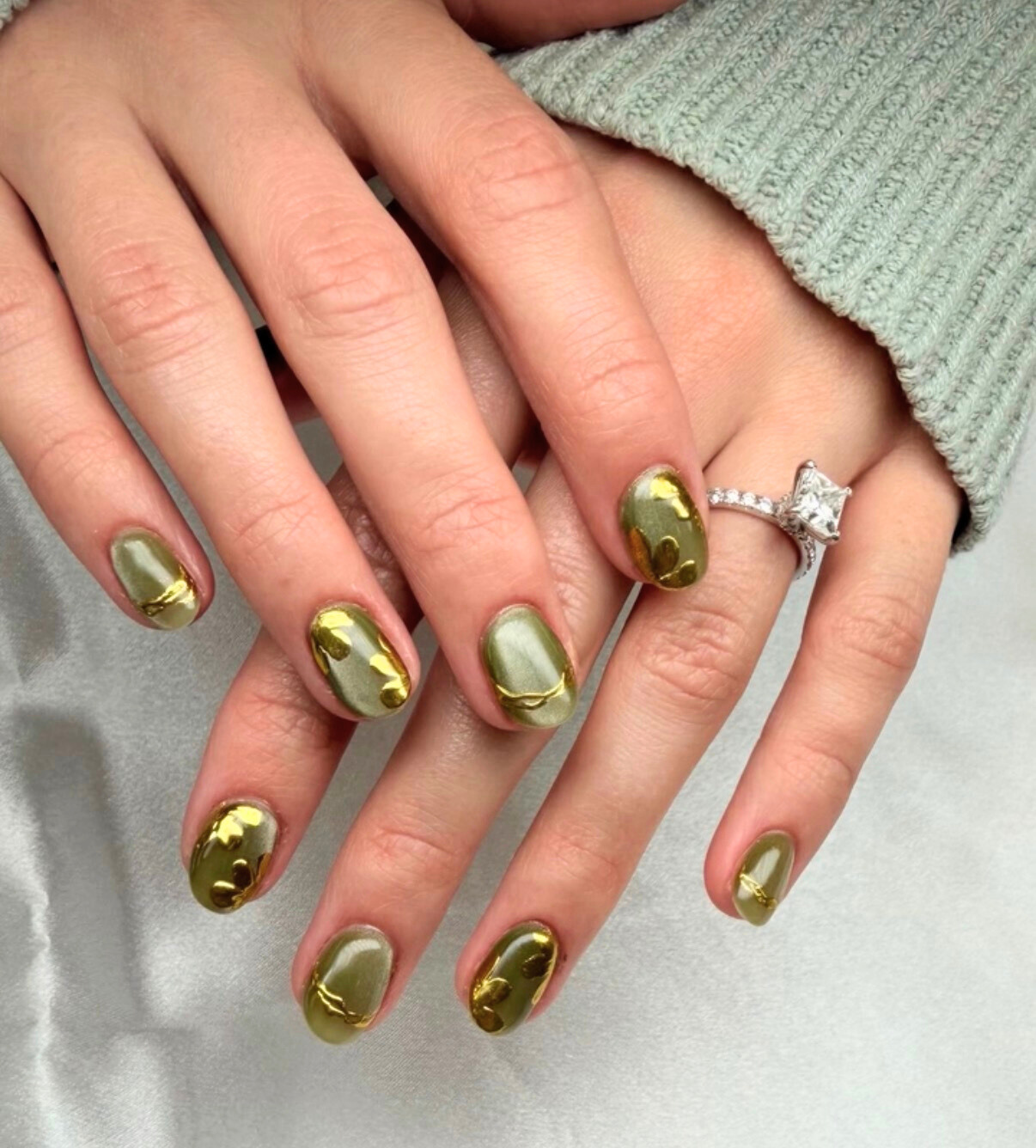 Close-up of hands with manicured nails painted in olive green with gold accents, wearing a silver engagement ring with a large square-cut diamond.