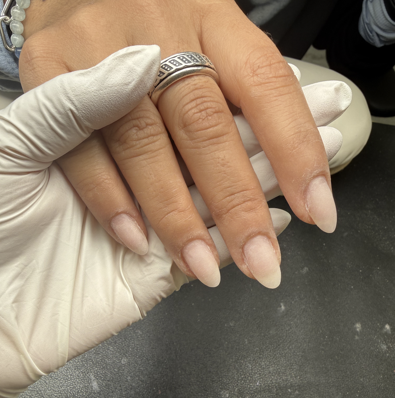 Close-up of a person's hand with long, almond-shaped, natural-looking nails being held by a gloved hand in a clinical setting.
