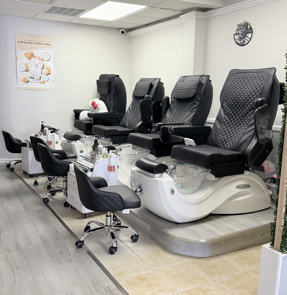 A salon pedicure station with three black reclining chairs and foot basins, along with small black chairs and manicure tables in front, in an indoor salon setting.