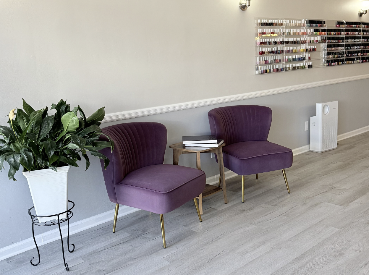 A waiting area with two purple velvet chairs with gold legs, a small wooden side table with books, a large green plant in a white pot on a black metal stand, gray walls with nail polish displays, and a white air purifier on the floor.