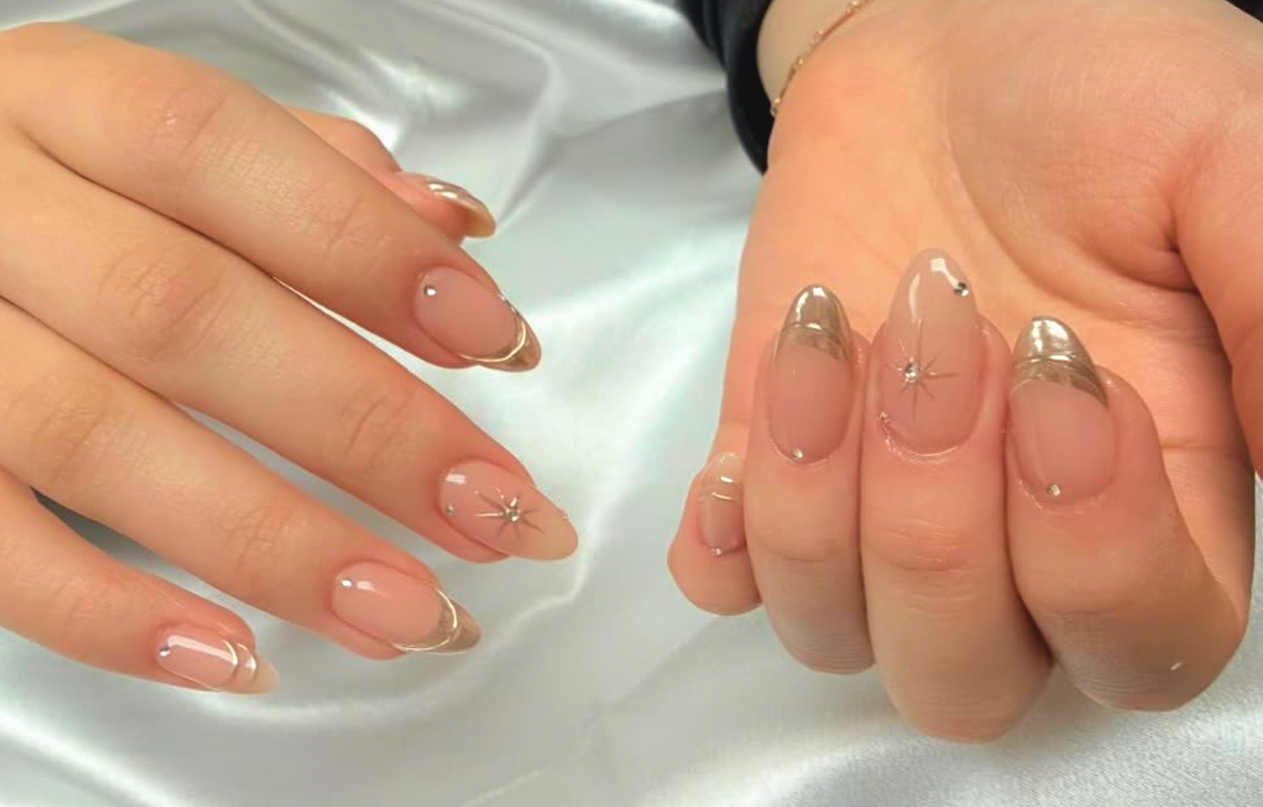 Close-up of a person's hands with manicured nails featuring a clear base coat, subtle gold accents, and small decorative details, resting on a silky surface.