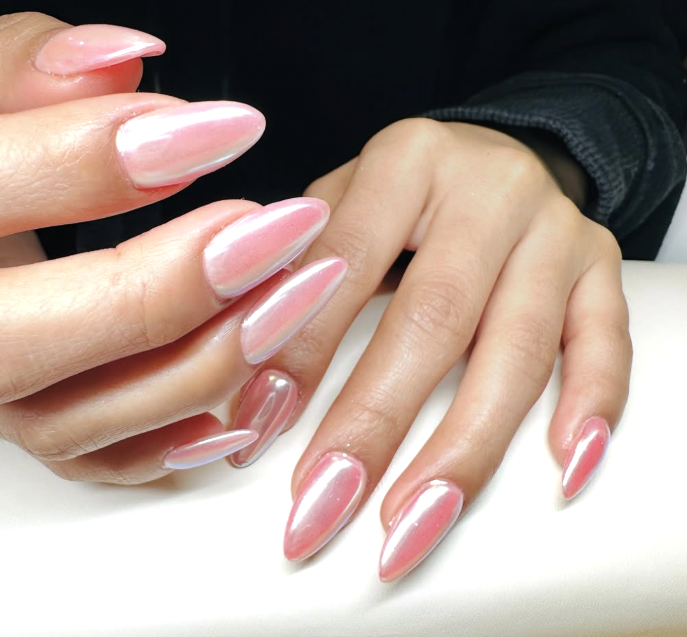 Close-up of a person's hand with neatly manicured nails painted in a shiny, iridescent pink color, resting on a white surface.