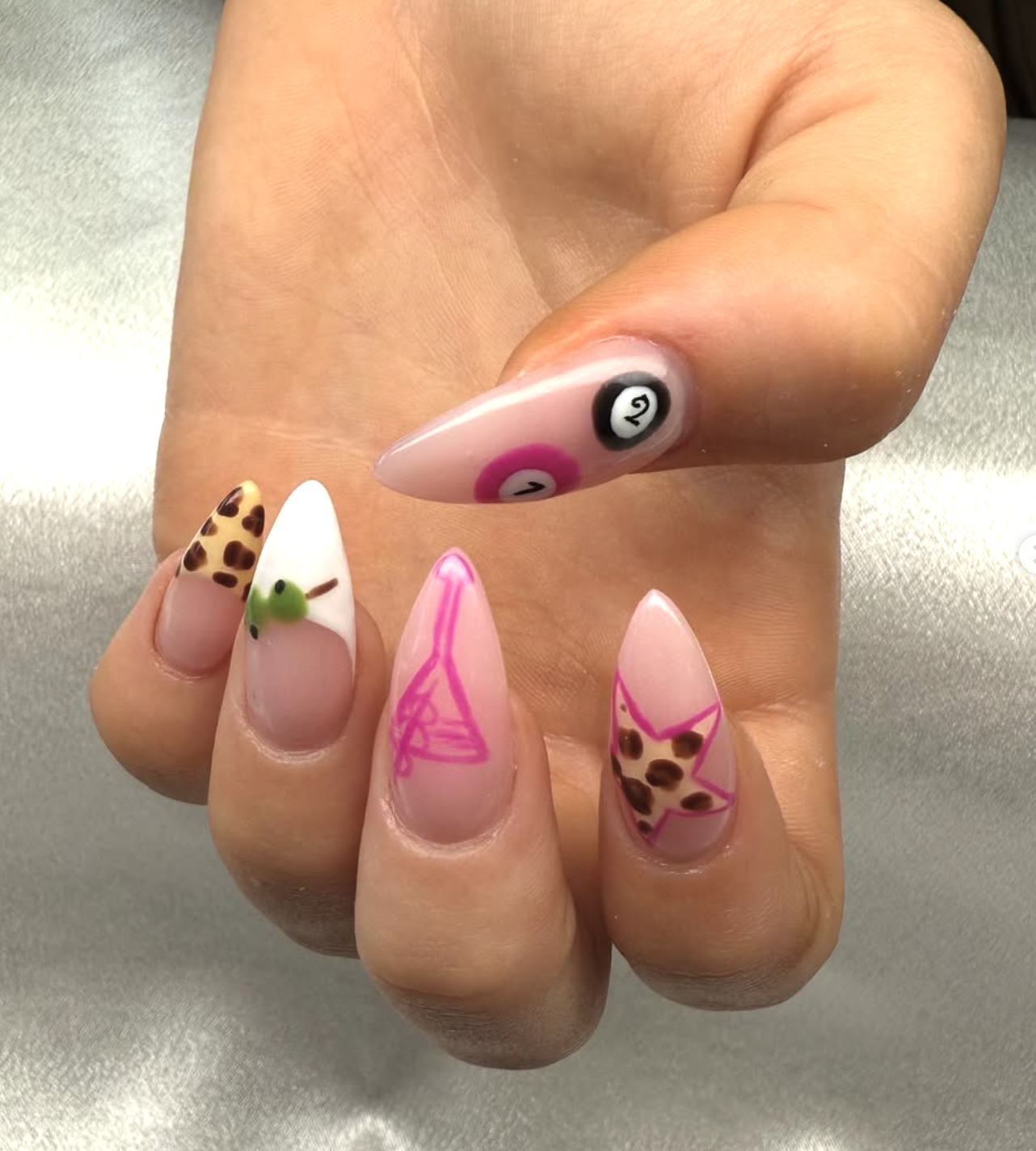 Close-up of a hand displaying five decorated nails with various designs, including animal print, a chick, pink line drawings, and intricate nail art.