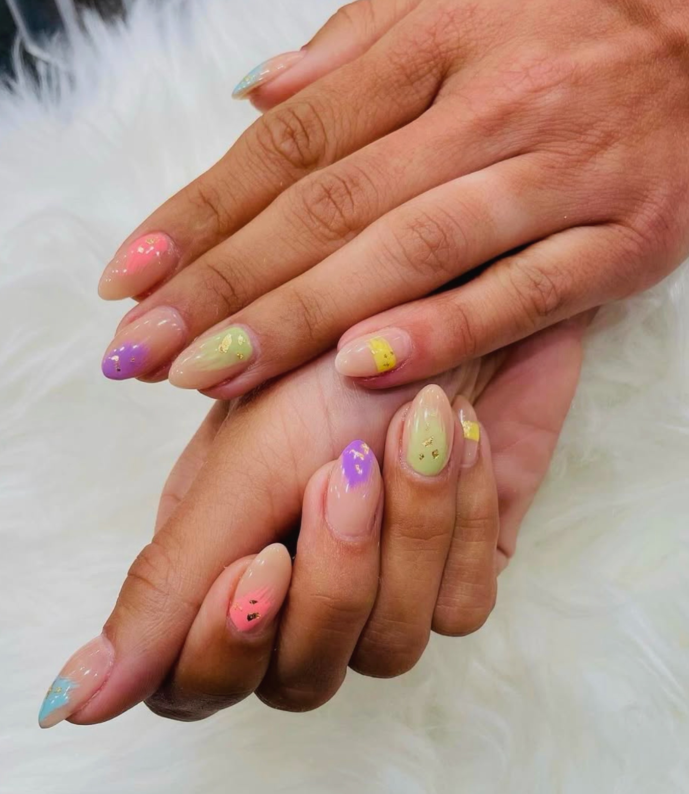 Close-up of two hands with pastel-colored nail polish and gold foil accents, resting on a white, fluffy background.