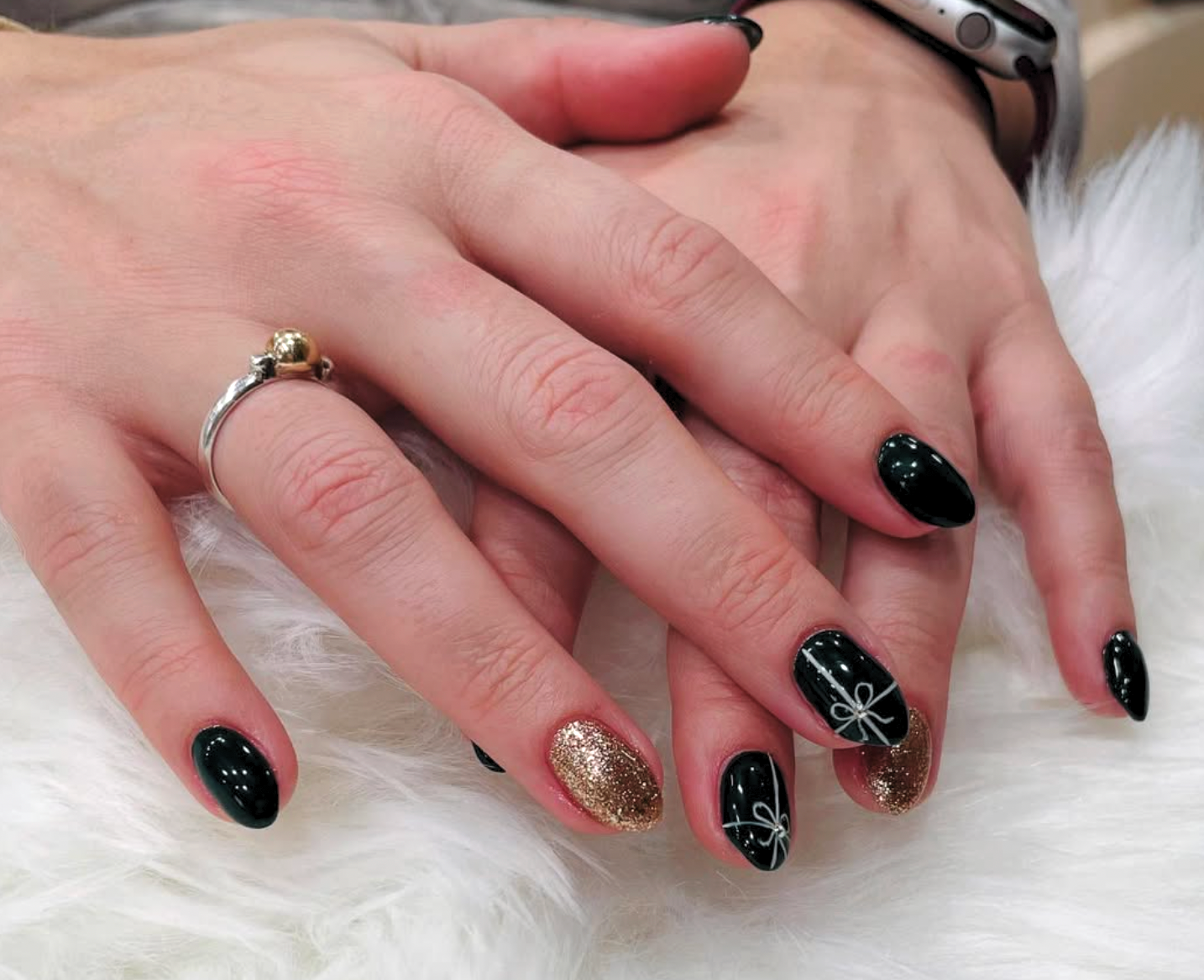 Close-up of a person's hands resting on a white furry surface, showing manicured nails with black, gold glitter, and white holiday-themed designs. The person is wearing a silver ring with a small gold sphere on the finger.