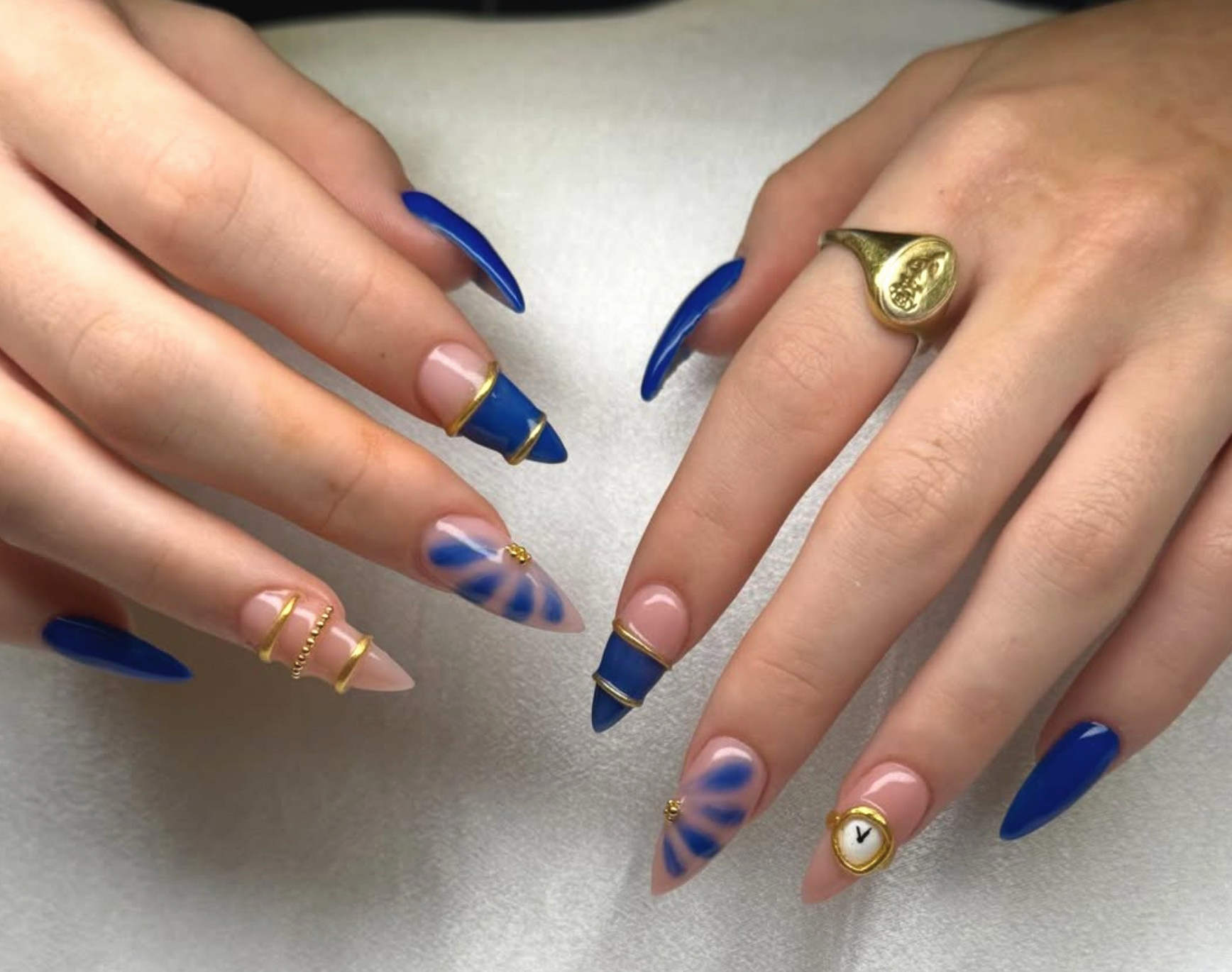 Close-up of hands with decorated nails and jewelry, featuring blue and pink nail polish designs, gold rings, some with chain details, and a watch with a white face and black hands.