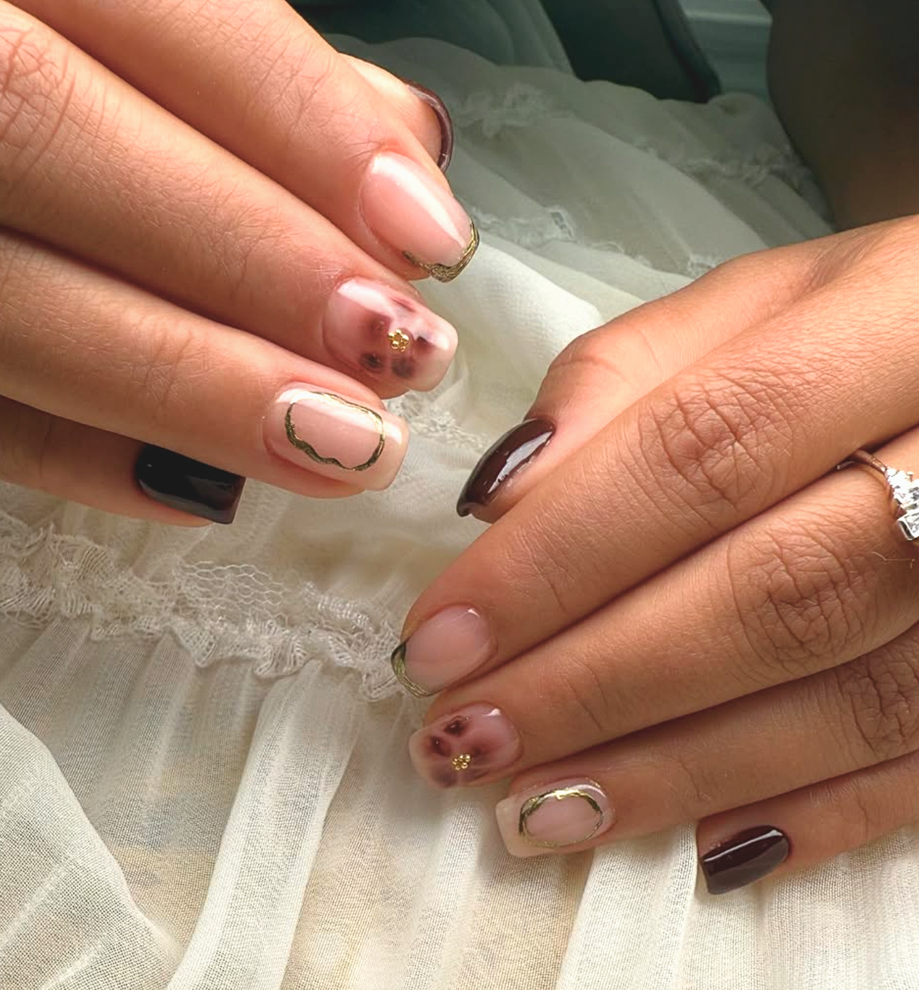 Close-up of hands with manicured nails resting on a white lace fabric. The nails feature various designs, including black, nude, and floral patterns with gold accents.