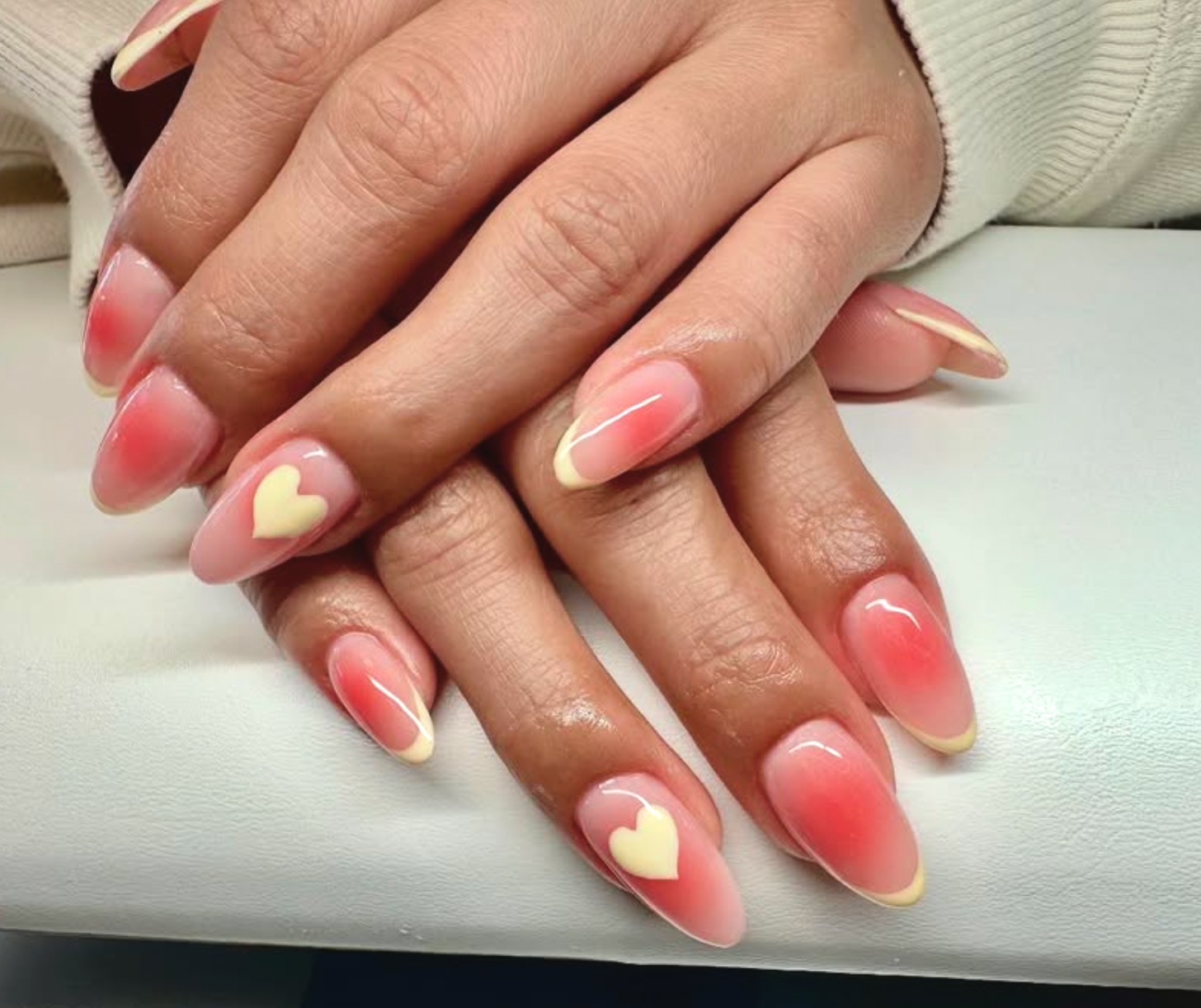 Close-up of hands with manicured nails featuring pink gradient polish and a white heart design on two nails.