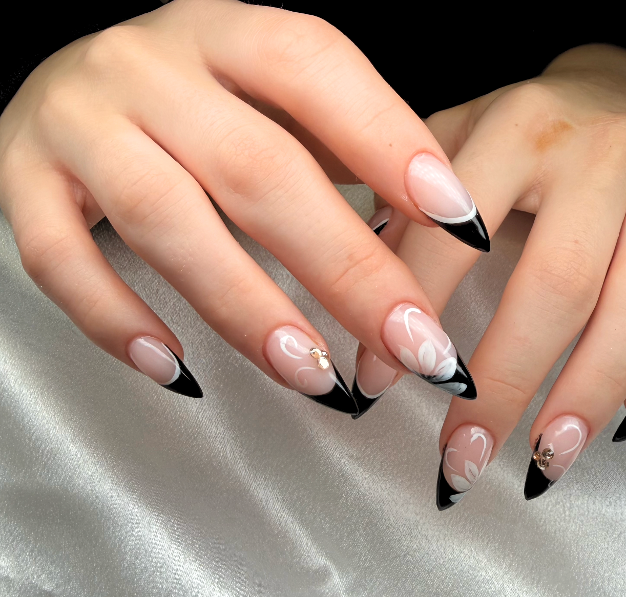 Close-up of hands with black and white French manicure nails, some with decorative rhinestones, resting on shiny silver fabric.