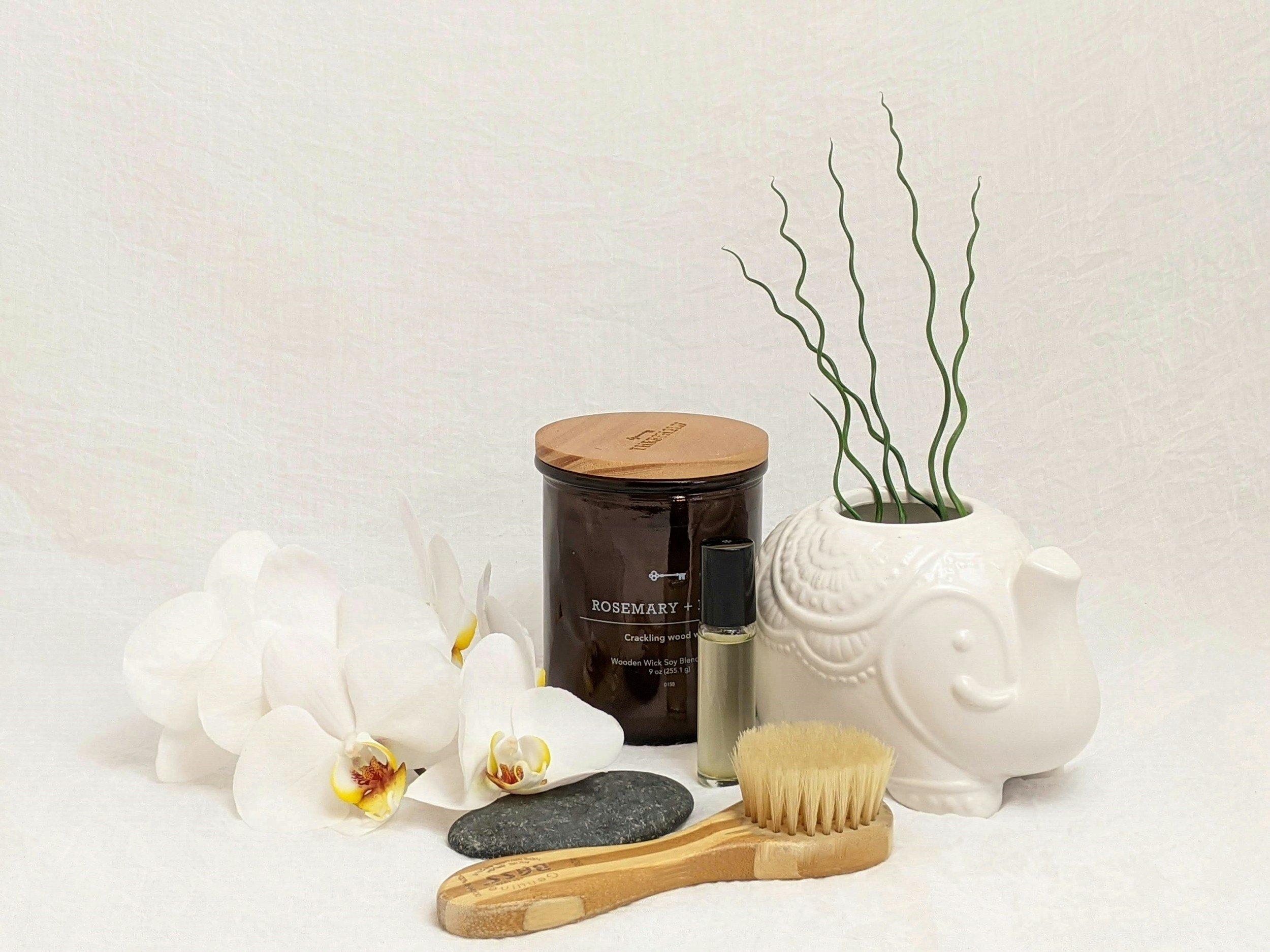 A collection of wellness and relaxation items including white orchids, a black stone, a wooden massage brush, a scented candle, a container labeled rosemary wood wick blend, a small essential oil bottle, a white decorative sculpture, a jar with a wooden lid, and a white orchid in a vessel, all arranged against a plain white background.