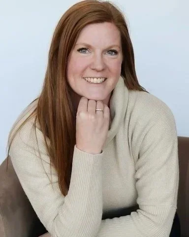 A woman with long red hair, smiling, wearing a beige sweater, resting her chin on her hand, sitting against a light background.