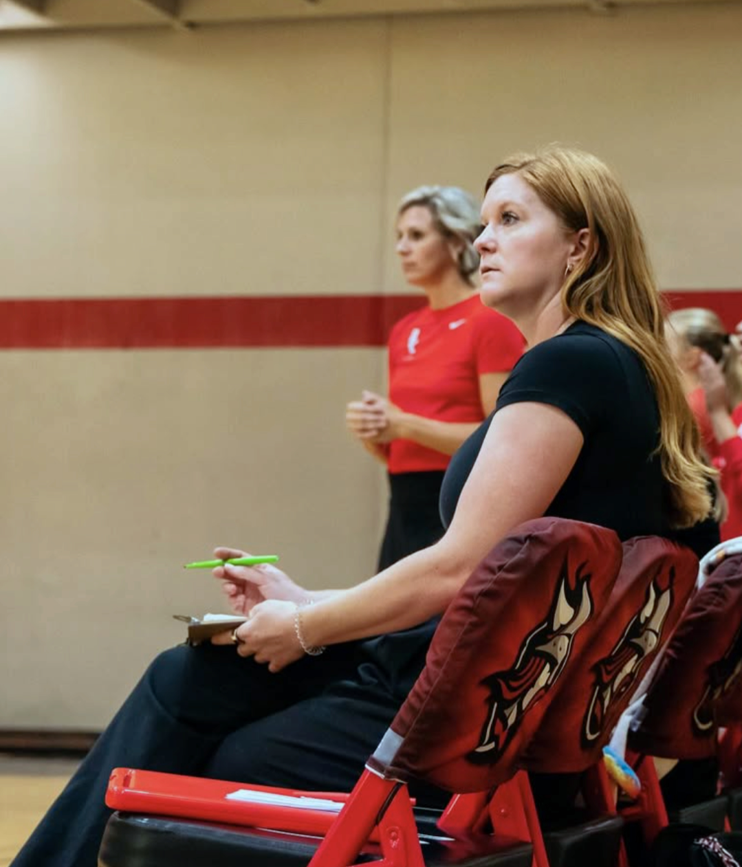 Two women seated and standing in a gymnasium, paying attention during a basketball game. The woman in the foreground has long red hair, is wearing a black shirt, and is holding a green pen and notebook. She is sitting on a red chair with a towel draped over the back. The woman in the background has short gray hair, is wearing a red shirt, and is standing with her hands clasped.