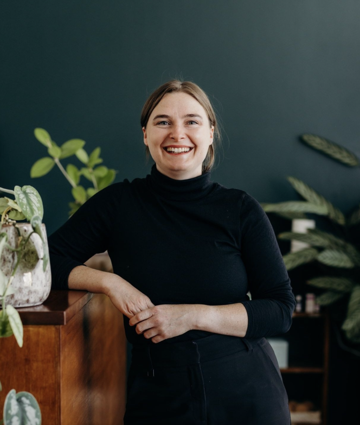 A young woman with light brown hair smiling and wearing a black turtleneck, standing indoors next to a wooden piece of furniture and green indoor plants, with a dark green wall in the background.