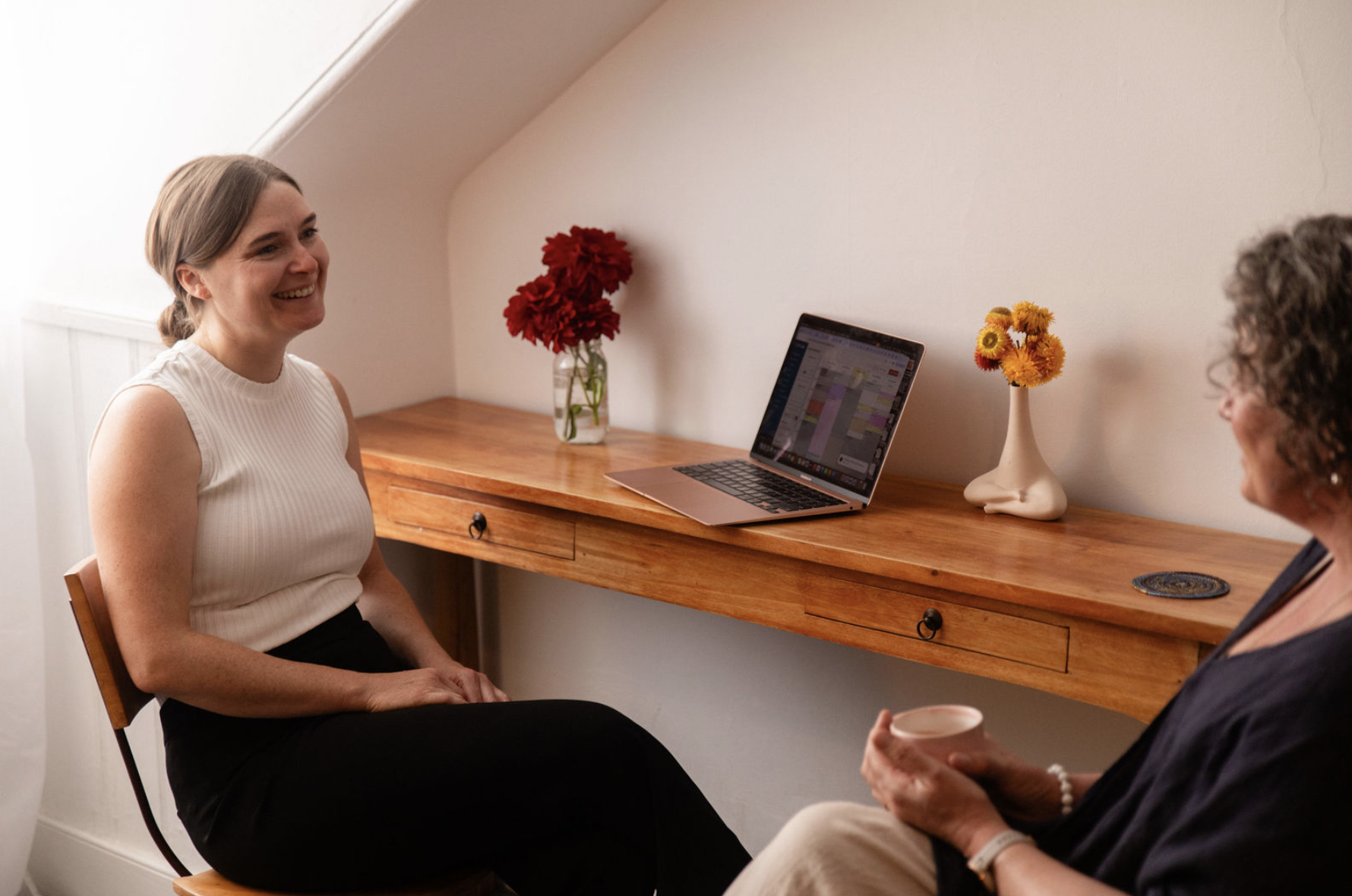 Two women seated facing each other in conversation in a cozy room with a wooden table and flowers, one woman smiling while the other holds a cup.
