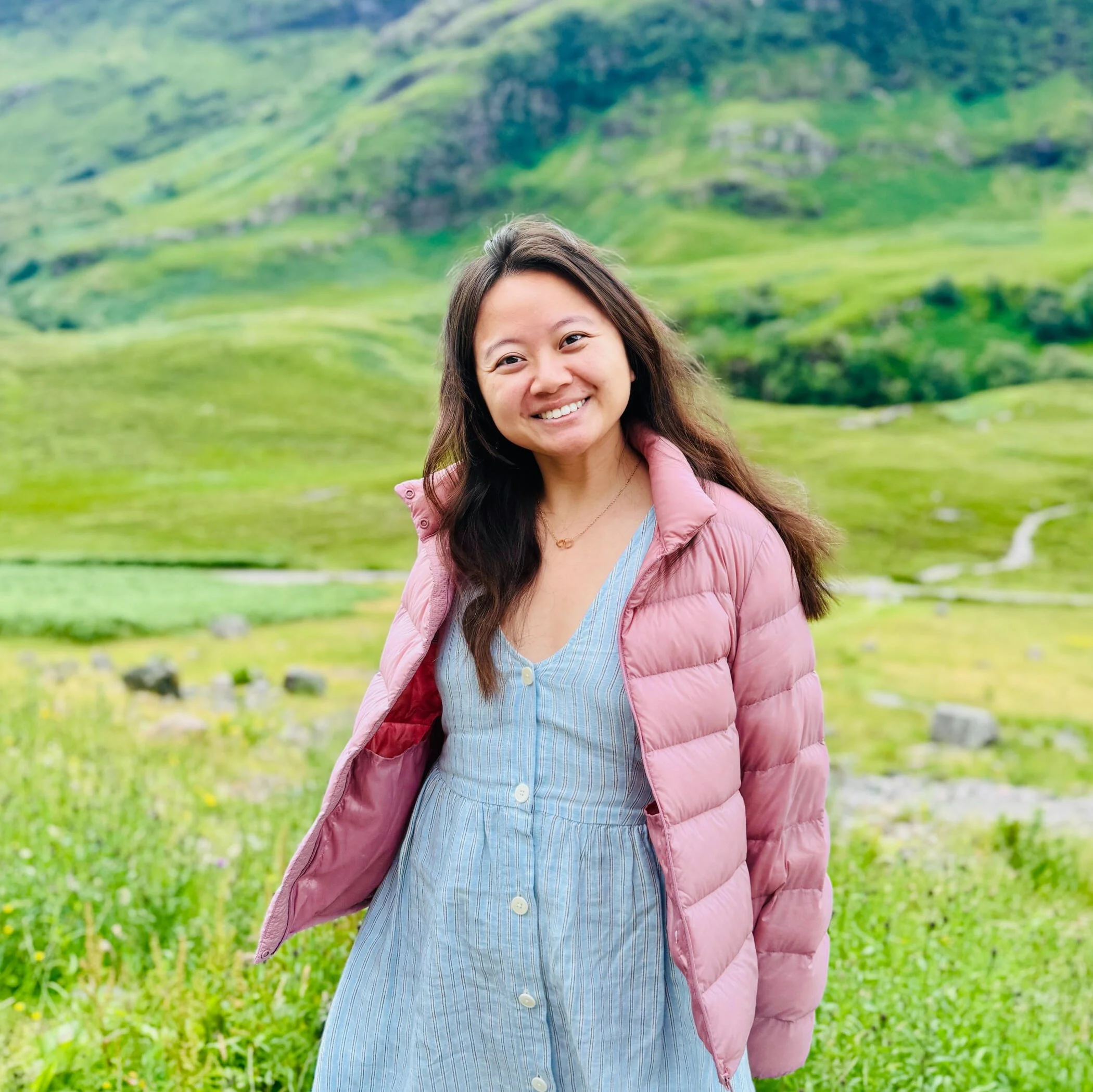 A young woman with long dark hair smiling outdoors, wearing a pink puffy jacket over a blue and white striped shirt, standing in front of green hills and grass.