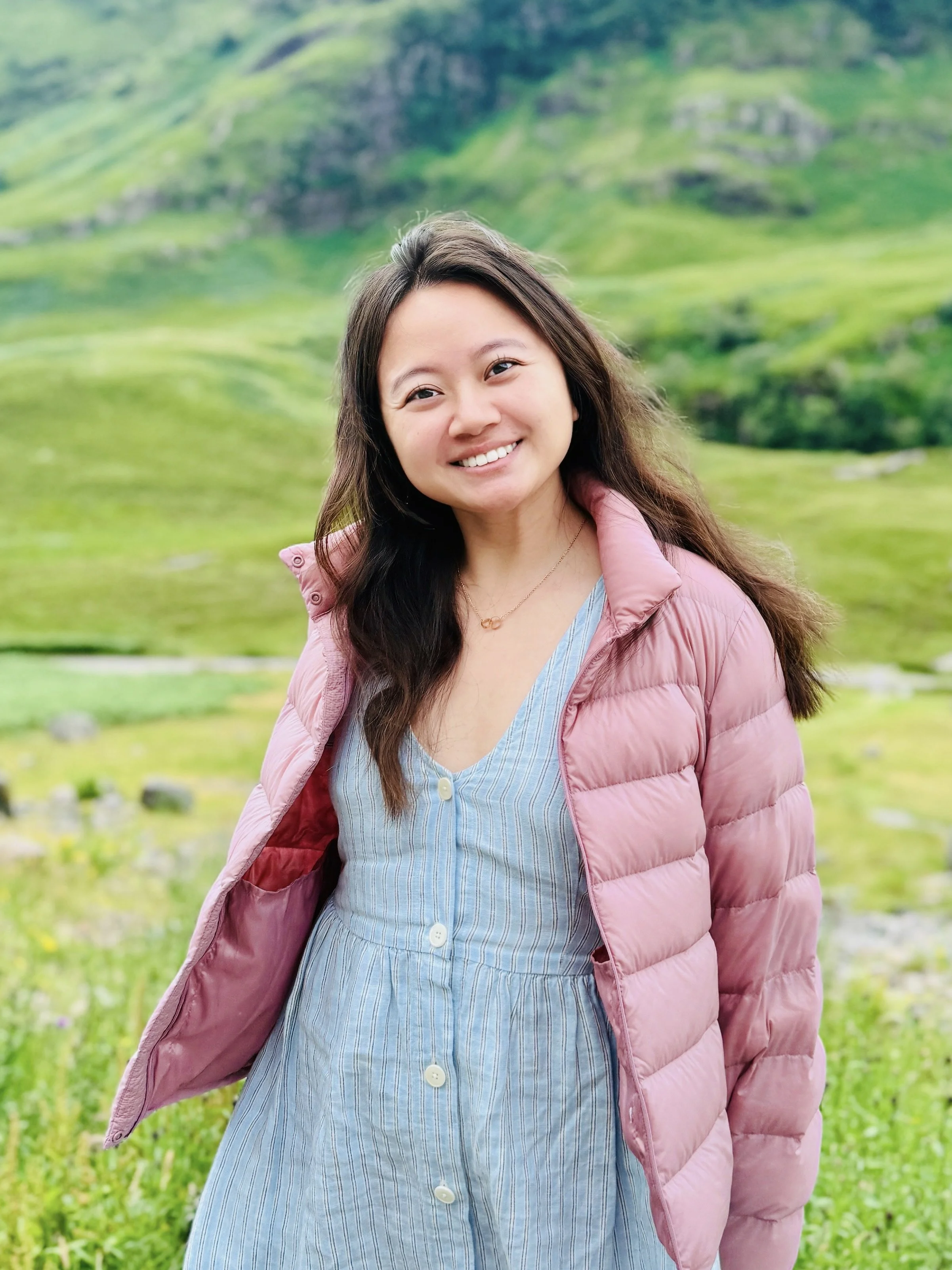 A woman with long dark hair smiling outdoors in a green mountainous area, wearing a pink puffer jacket over a blue and white striped dress.