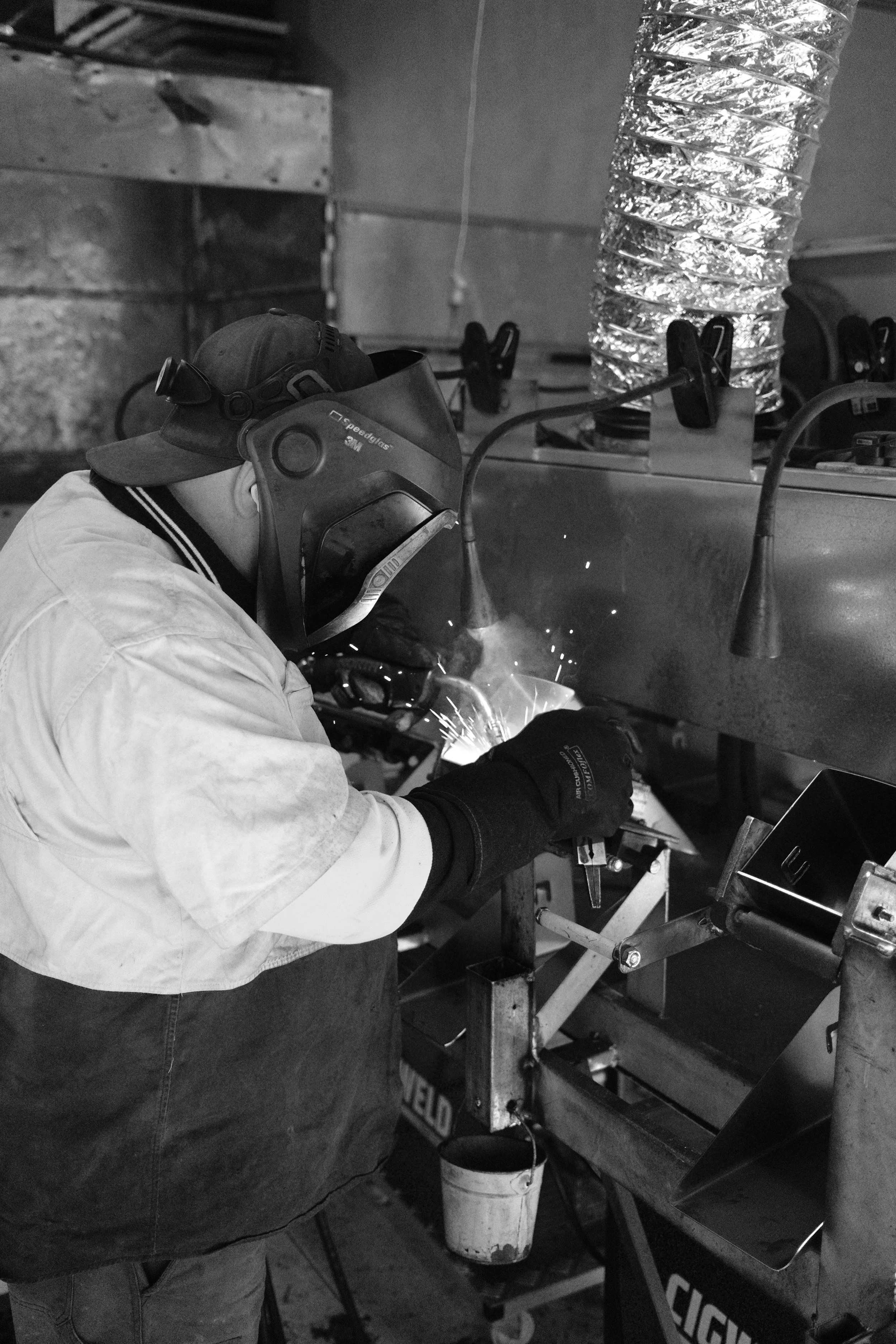 A person wearing protective gear, including a welding mask, welding gloves, and safety glasses, is welding an AdaptaKerb in a workshop.