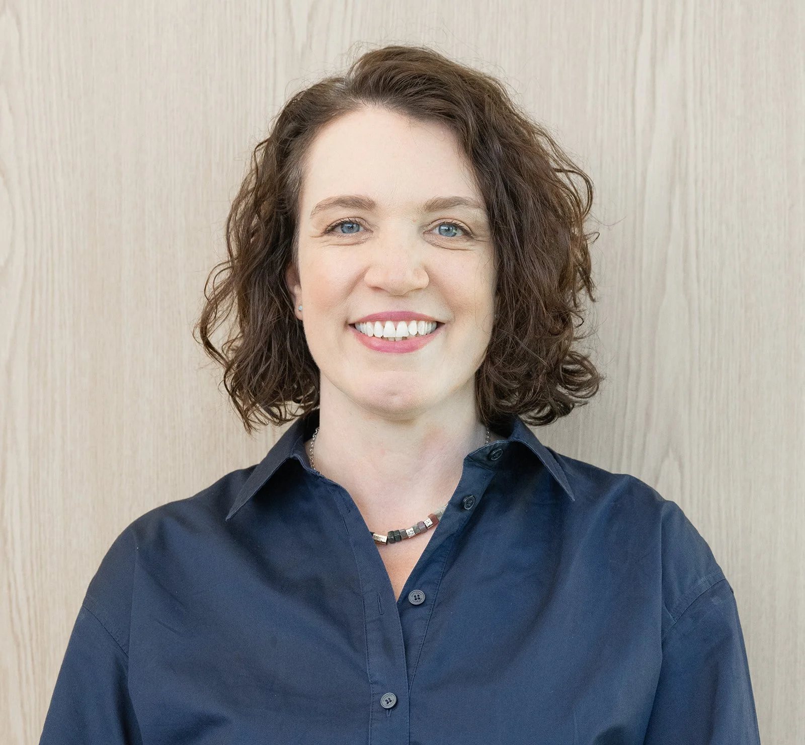 A woman with curly brown hair, light skin, and blue eyes smiling, wearing a black shirt and a beaded necklace, standing against a light-colored wooden background.