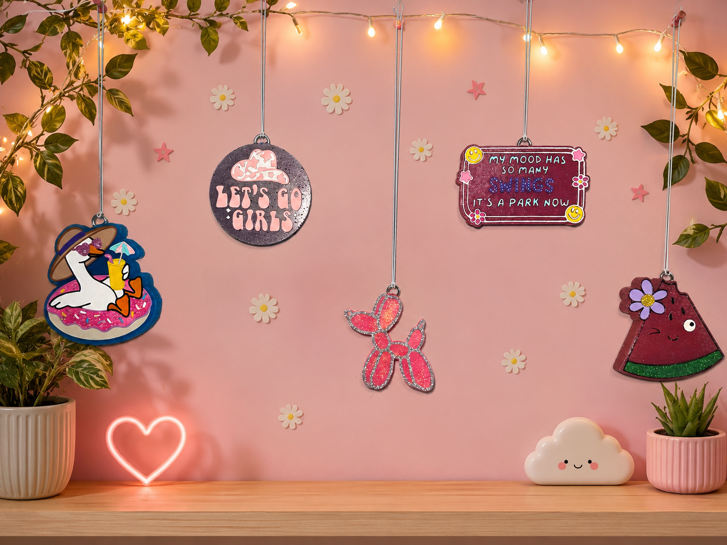 Decorated pink wall with hanging ornaments, string lights, potted plants, a neon heart, and a cloud-shaped light on a wooden surface.
