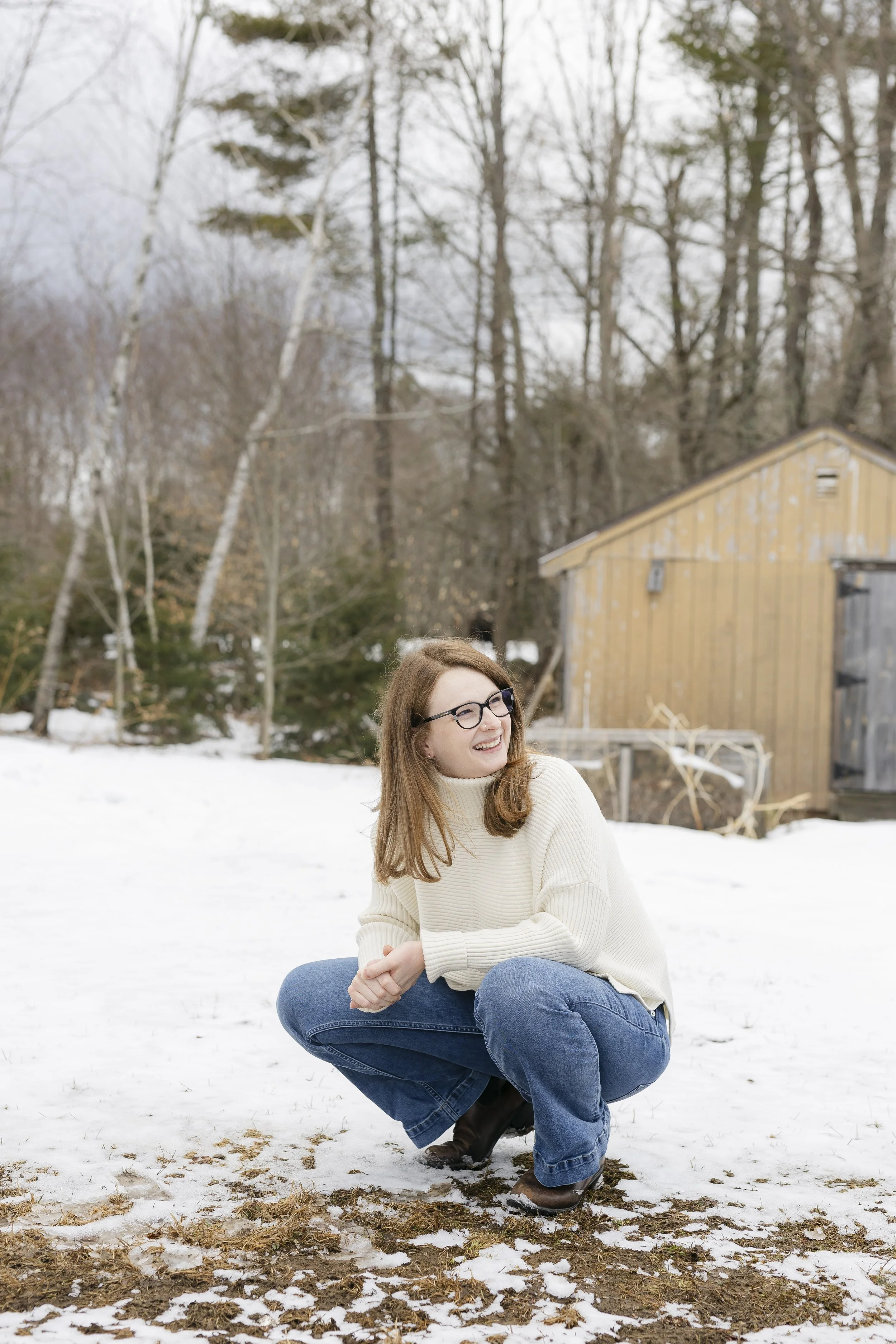 A woman with glasses and long hair squatting outdoors in the snow, wearing a cream sweater, blue jeans, and brown boots, smiling and looking to the side. Behind her are leafless trees, a wooden shed, and patches of snow on the ground.