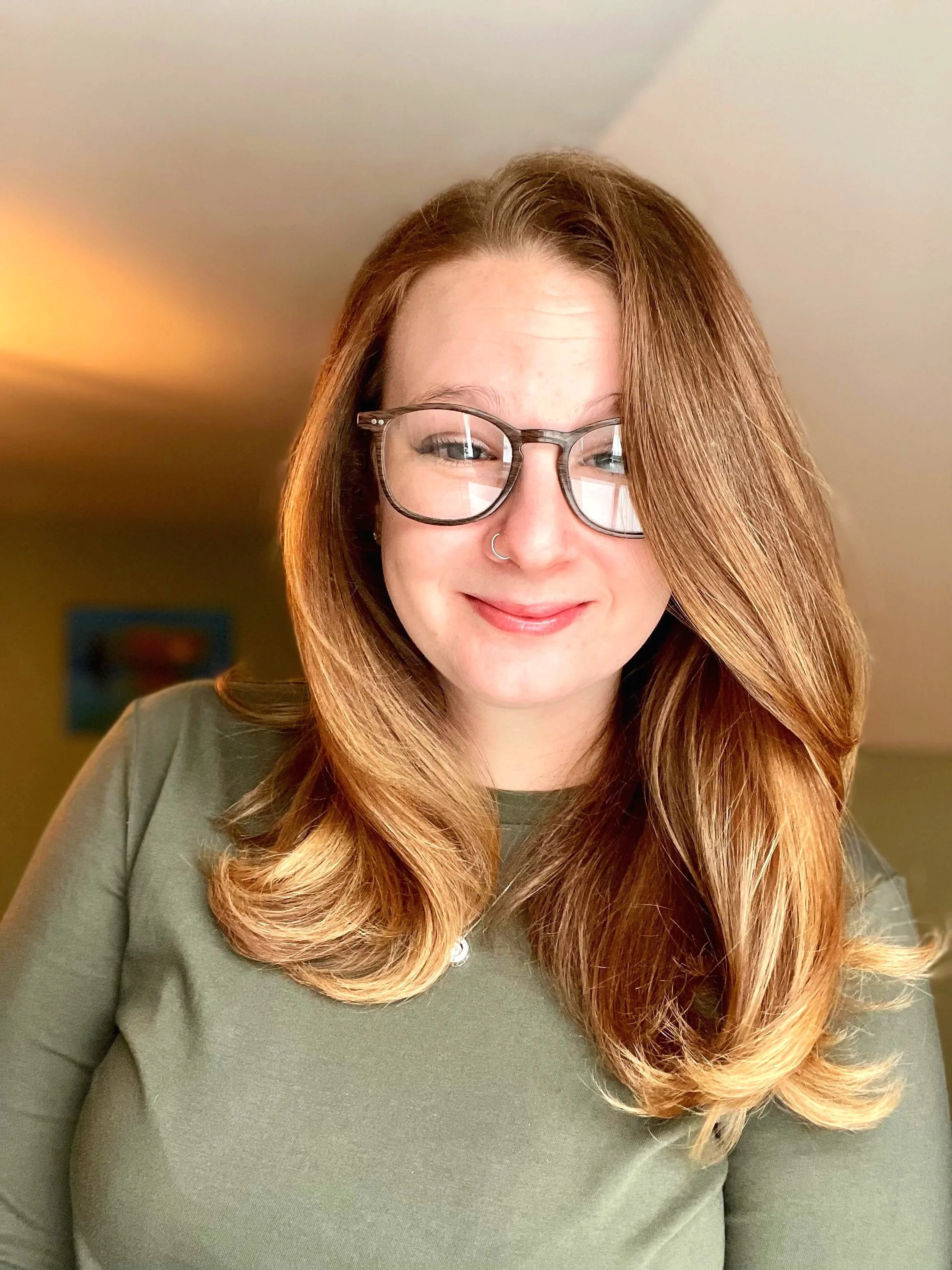 A woman with red hair, glasses, and a nose ring, smiling and wearing a green top, in a room with a yellow wall and a blue picture in the background.