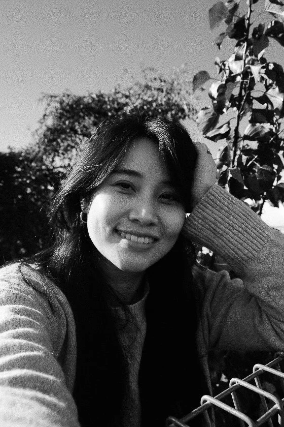 A photo of founder of Hearth, Helena Willows smiling towards the camera in black and white, surrounded by trees and sunshine.