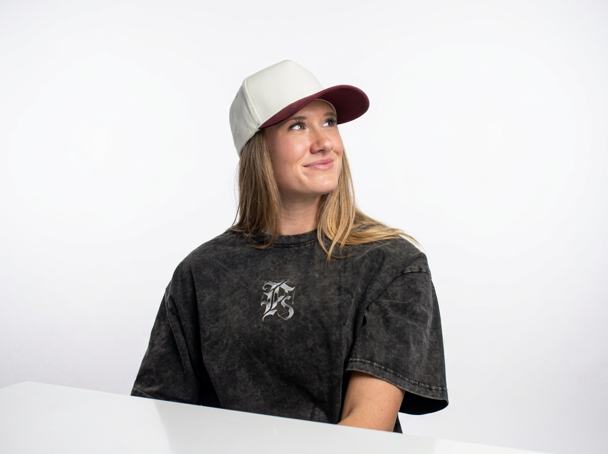 A young woman with long blonde hair wearing a beige and red baseball cap and a black T-shirt with a Gothic 'F' logo, sitting against a white background and looking to her left with a slight smile.