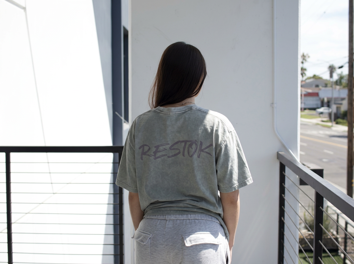 Woman standing on a balcony wearing a gray T-shirt with 'RESTOK' written on the back and light gray shorts, facing away from the camera.