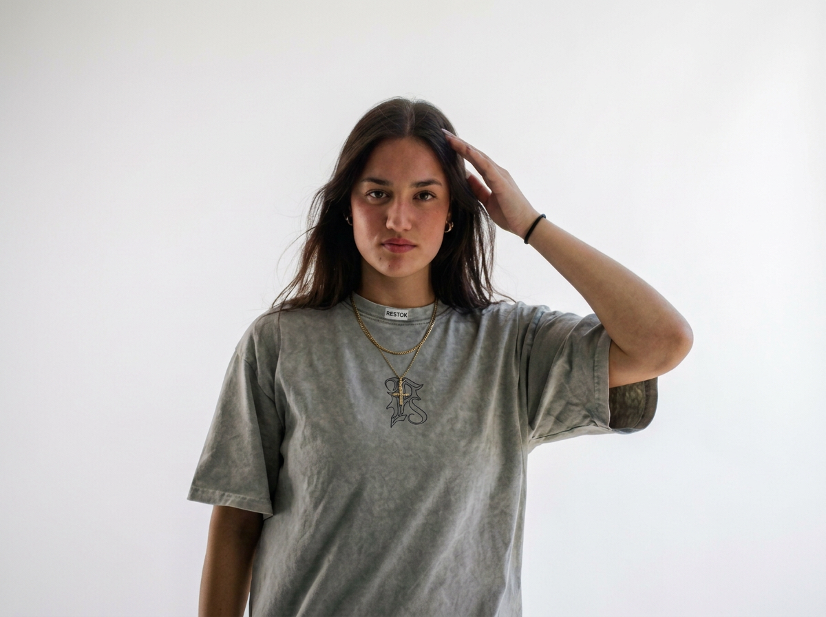 A young woman with long brown hair wearing a gray T-shirt with a graphic, layered necklaces, and a black bracelet, standing against a plain white background, touching her hair with her right hand, looking directly at the camera.