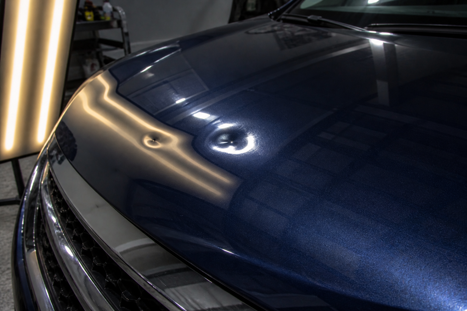 Close-up of the front hood of a shiny, dark blue luxury car with a sleek grill, reflecting overhead lights in a garage.