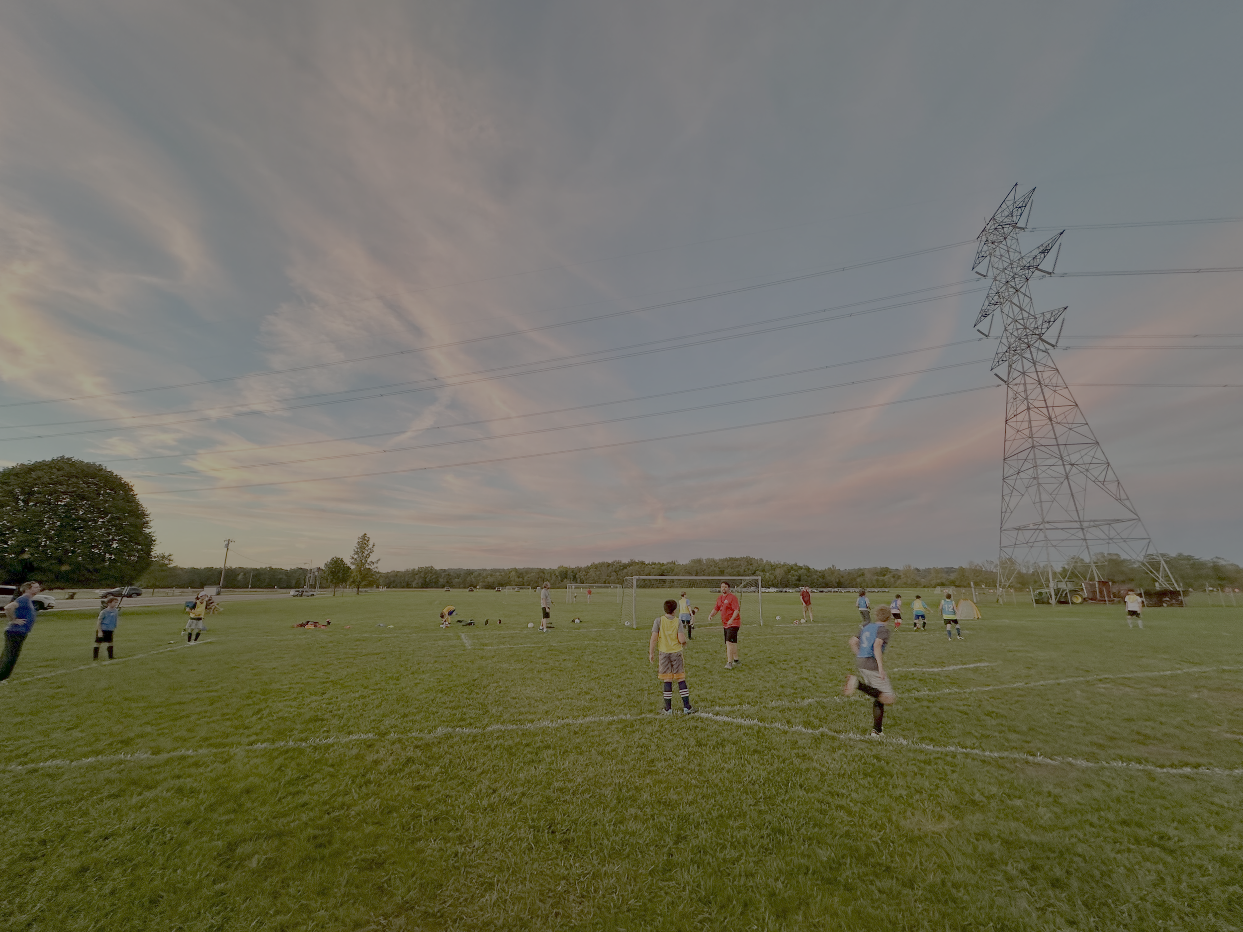 Soccer game with children and adults playing on a grass field under a cloudy sky, with a large electrical transmission tower on the right and trees in the background.