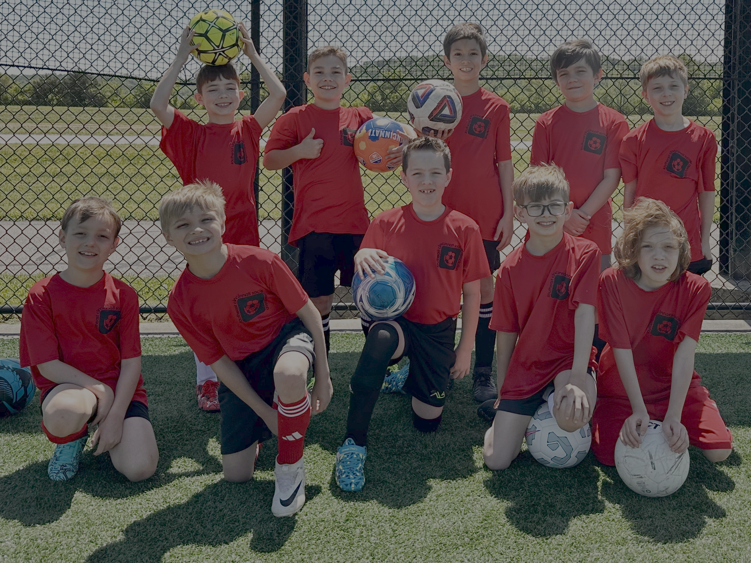 A group of ten young boys wearing red soccer jerseys, posing on a soccer field with soccer balls, in front of a chain-link fence. Some are kneeling, others standing, smiling at the camera.