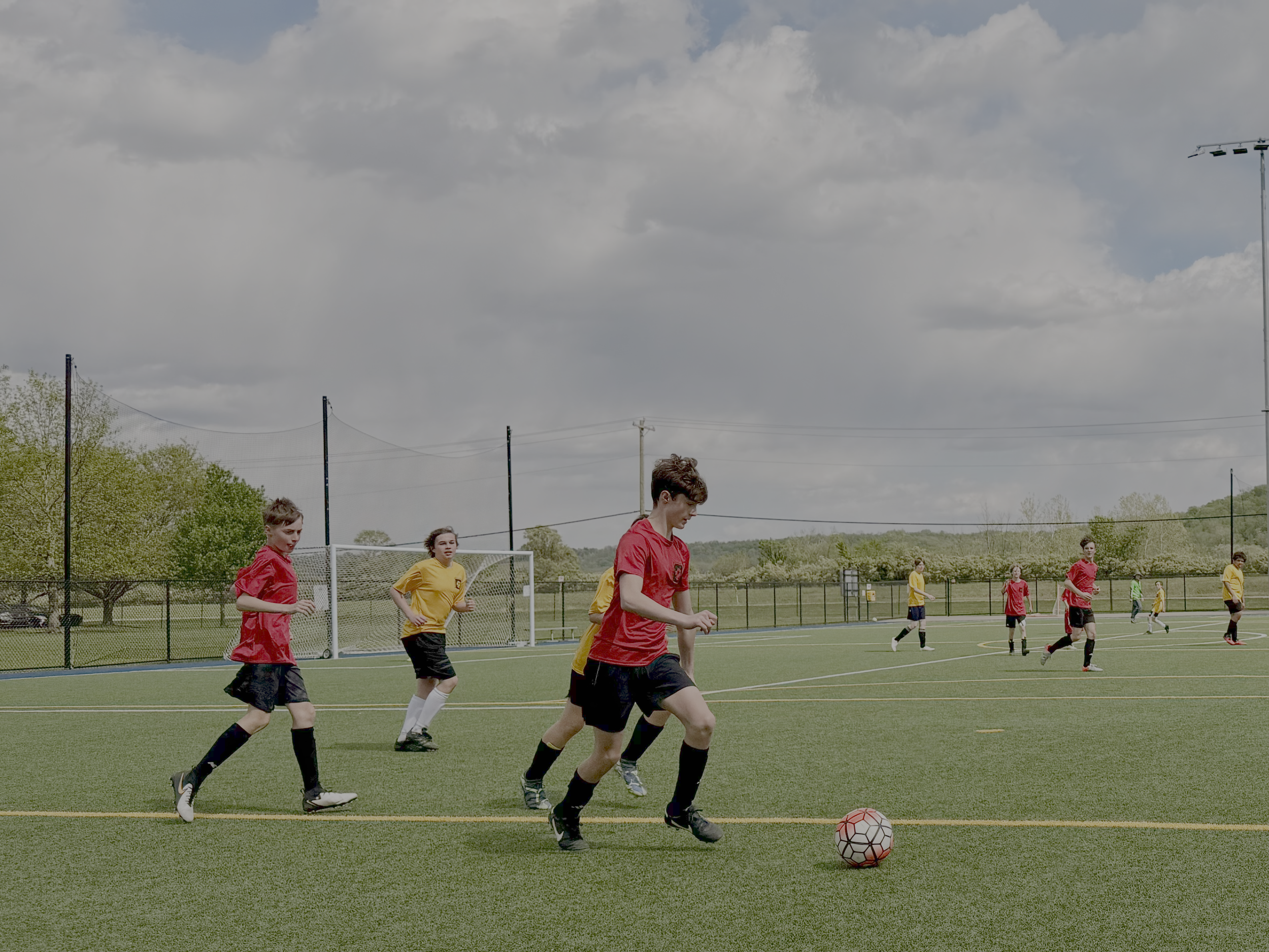 Young children playing soccer on a field under cloudy sky, wearing red and yellow jerseys.