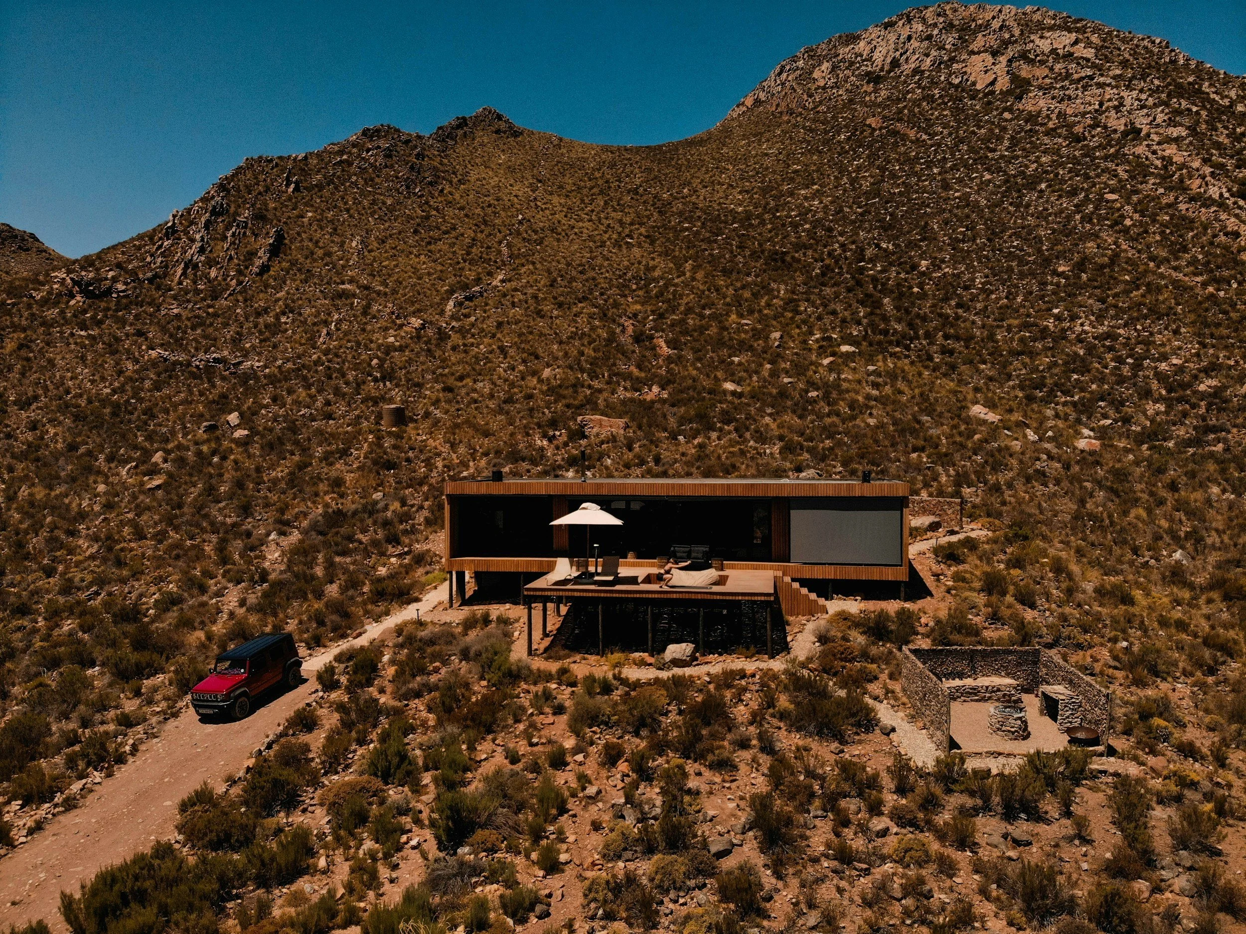 Modern house with large glass windows and patio furniture, situated at the base of a rocky mountain under a clear blue sky.