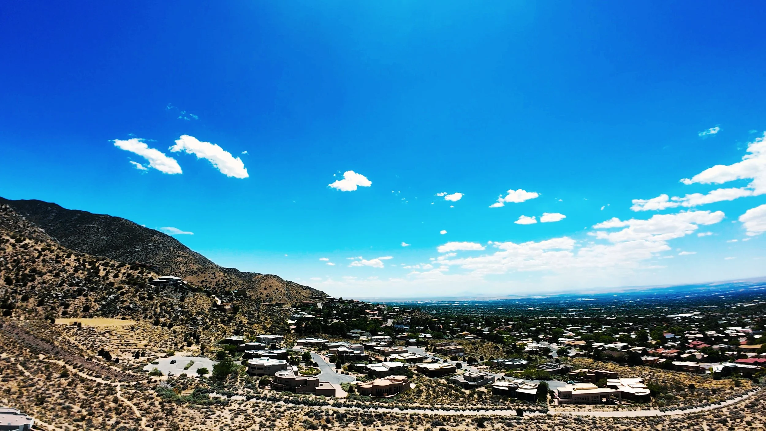 A landscape view showing a hillside residential area with houses, and a cityscape in the distance under a bright blue sky with scattered clouds.