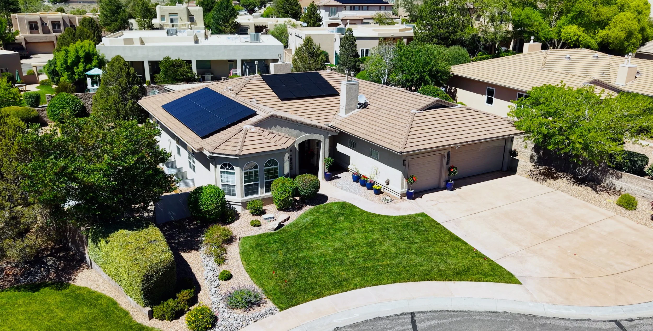 A house with solar panels on the roof, surrounded by a landscaped yard with green grass, bushes, and trees. The house has a driveway and a two-car garage, with potted plants along the front.