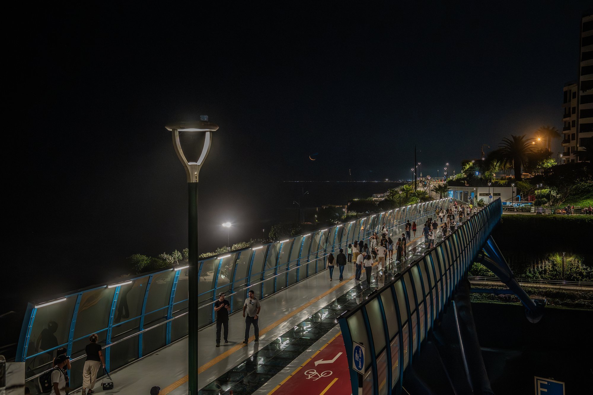 Puente peatonal iluminado de noche con gente caminando junto al mar