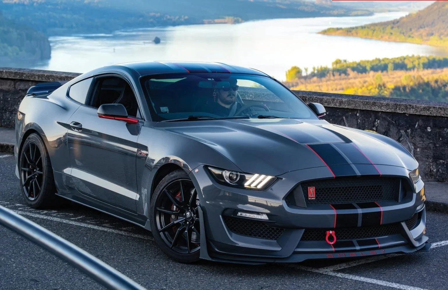 Grey Shelby sports car with red and black racing stripes parked by a scenic overlook with a river and hills in the background.