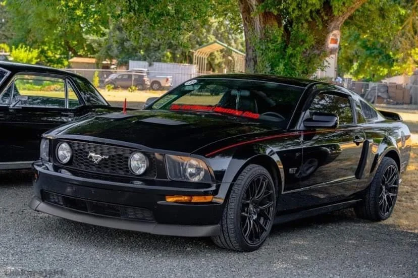 Black Ford Mustang GT parked outdoors on gravel, with trees and other vehicles in the background.